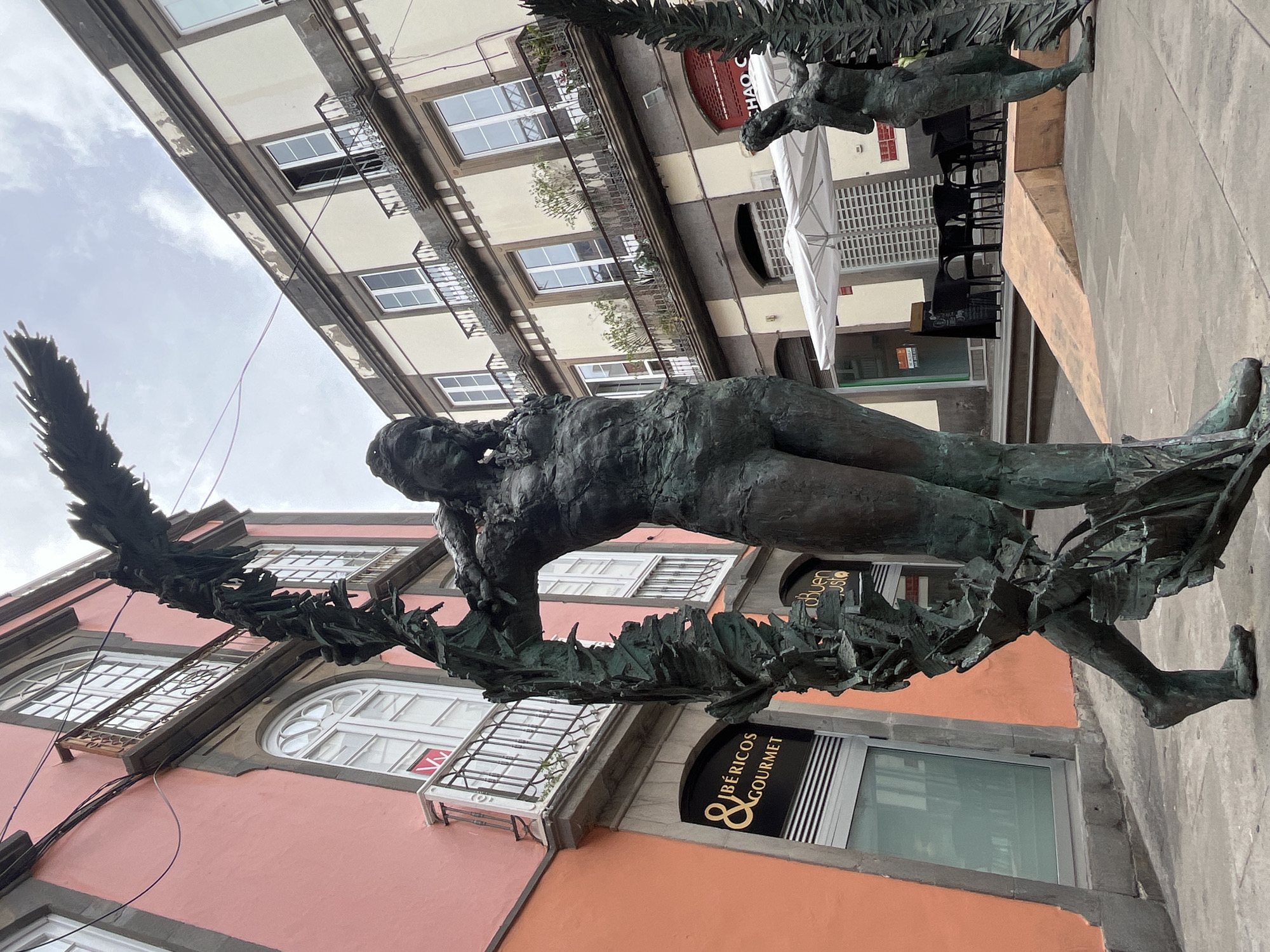 The image shows a bronze statue in Las Palmas De Gran Canaria, Spain. The statue depicts a nude, reclining figure resting on a leafy branch or wreath. The statue is located in front of buildings. One building is peach-colored and features arched windows and a sign that reads "Ibéricos Gourmet". Another building is white with multiple rectangular windows, some with small balconies. The sky is overcast. A few tables and chairs are visible in the background.