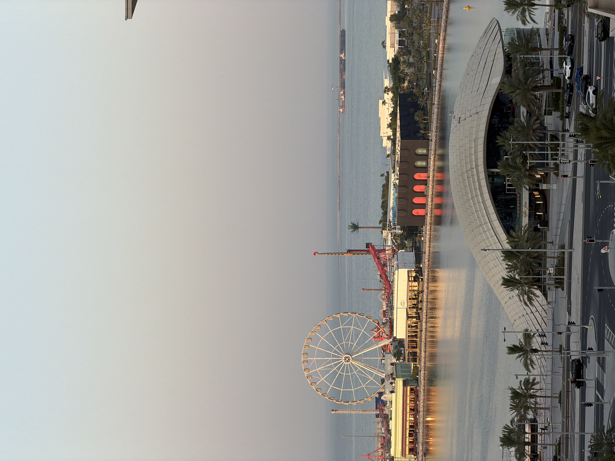 The image shows a waterfront scene with a Ferris wheel, buildings, palm trees, and water. The Ferris wheel is large and white, with many gondolas. It is located in an amusement park area, with red cranes and other rides visible in the background. Several modern buildings line the waterfront, some with distinctive architectural features. The water is calm and reflects the light from the setting sun, creating a warm glow. Palm trees are scattered throughout the scene, adding to the tropical atmosphere. There are a few cars visible on a road to the right of the scene, and some people can be seen walking along the waterfront. The sky is a pale blue, and the overall impression is one of a vibrant and lively place. The time of day appears to be late afternoon or early evening, as indicated by the warm light and long shadows. The weather seems to be clear and pleasant. There is some Arabic text visible on a white sign. The scene suggests a tourist destination or a recreational area.