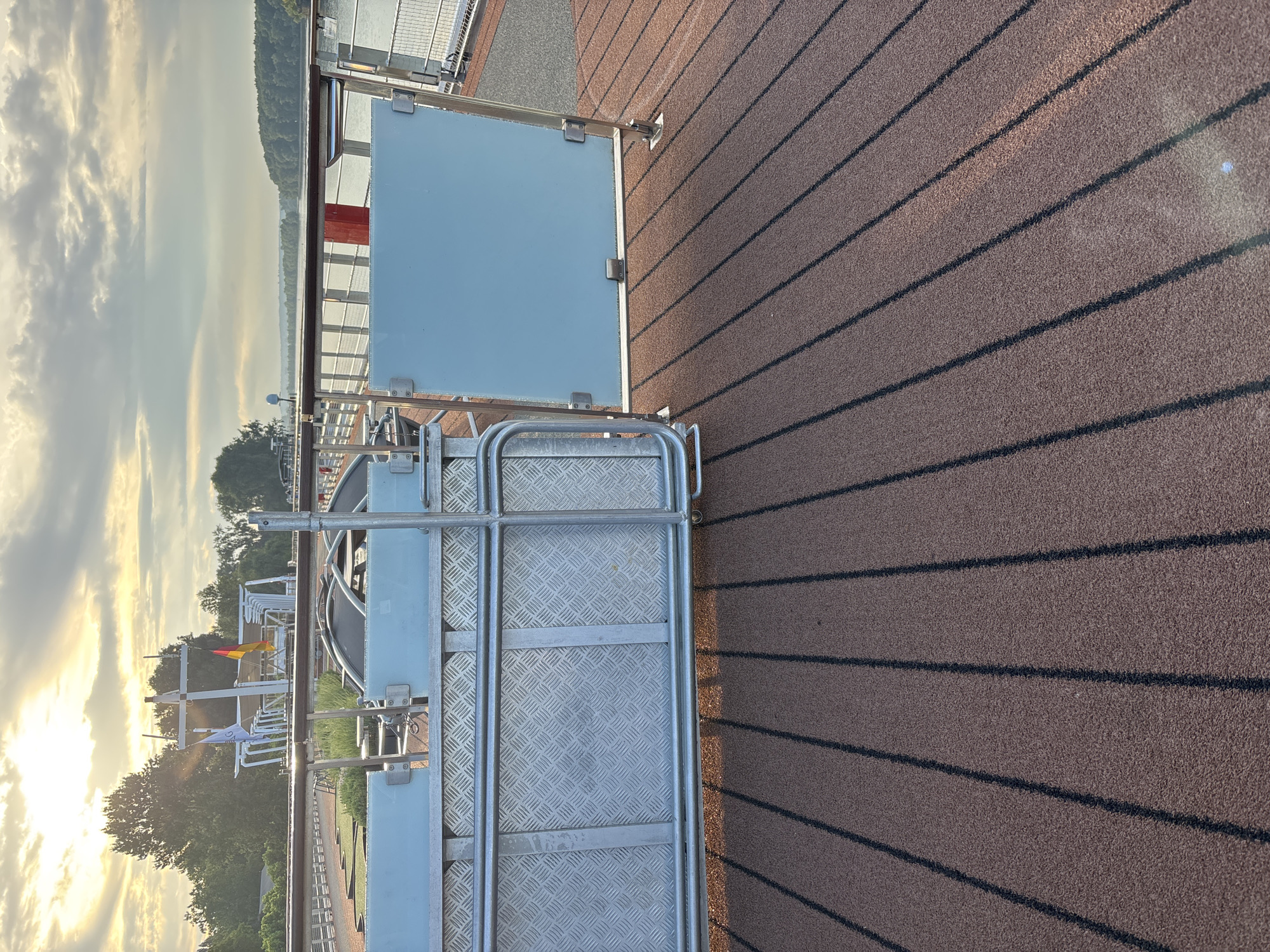 The image depicts a section of a deck or walkway, likely on a vessel or elevated structure, with a textured brown surface marked by dark parallel lines. In the foreground, a metal railing and a grated metal panel are visible, possibly part of a gate or access point. To the right, a light blue panel is also part of a barrier. In the background, a body of water stretches towards a tree-lined horizon under a cloudy sky with the sun breaking through. A white structure with flags, one of which is the German flag, is situated further in the distance. The setting appears to be outdoors, with natural light indicating it is daytime. The location is identified as Rüdesheim, Germany.