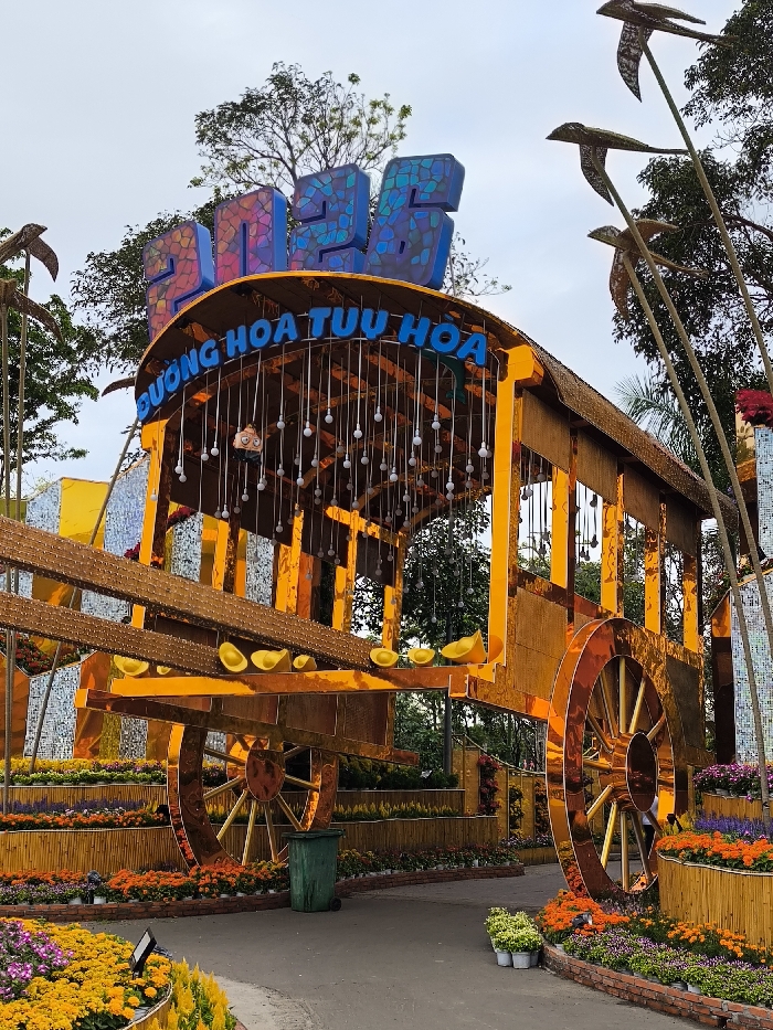 The image depicts a festive display on a flower street in Tuy Hòa City, Vietnam. A large, ornate golden-hued structure resembling an oversized antique wagon or cart dominates the central frame. Above its curved roof, a sign reads "2025" in multi-colored, textured block letters, with "ĐƯỜNG HOA TUY HÒA" in blue, illuminated letters positioned beneath it. Numerous white, spherical light bulbs hang suspended from the structure's interior roof, while several golden sycee (ingots) are placed along its horizontal elements. Tall poles adorned with decorative elements resembling flying creatures flank the wagon structure. The foreground and surrounding areas feature tiered flower beds densely planted with various colorful blossoms, including orange, yellow, and purple flowers. A dark paved pathway runs through the lower center, where a green plastic refuse bin is visible. A small cluster of potted plants sits on the path to the right of the bin. One individual is partially visible standing behind the right wheel of the wagon structure. The background consists of green trees and foliage under an overcast sky.