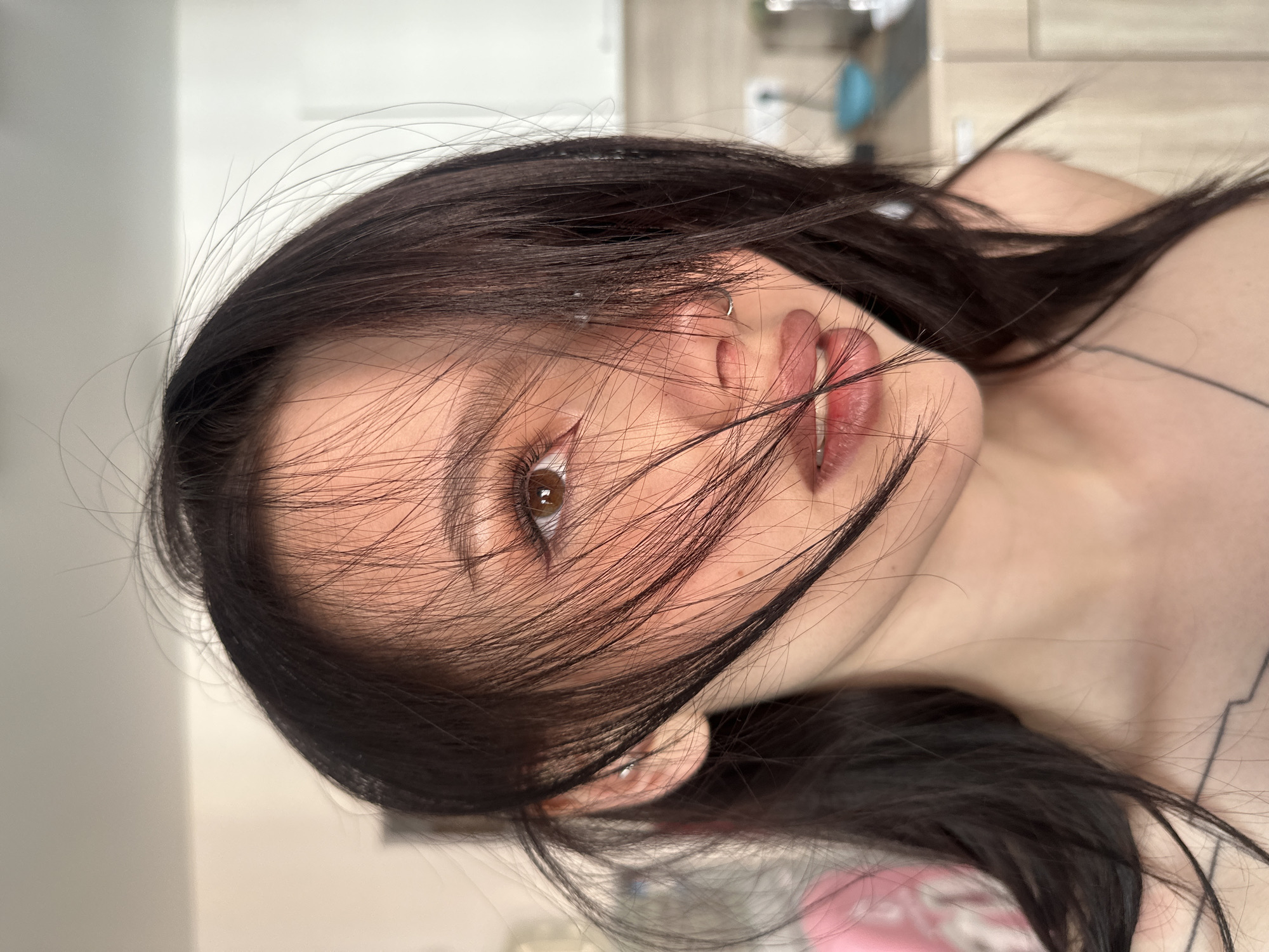 A close-up, portrait-oriented photograph depicts a young woman with dark hair partially obscuring her face. Her skin tone is fair. Her right eye, the only one fully visible, is brown. She has a septum piercing and an ear piercing. Her lips are partially open, revealing the upper row of teeth. The background is blurred and indistinct, showing elements suggesting a room interior. There is no contextual information available to determine the location in Lyubertsy, Russia.