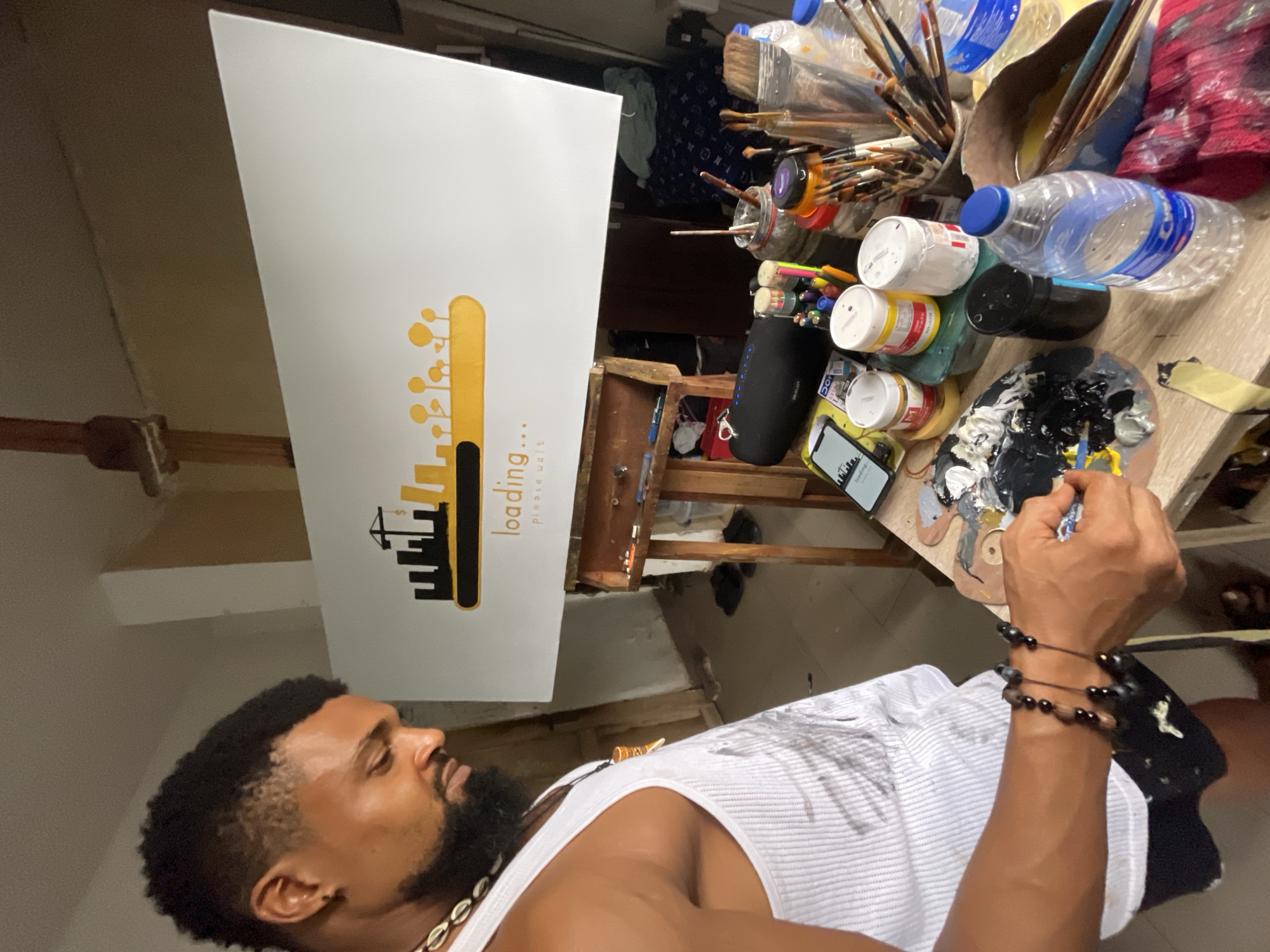 The image presents a man with dark skin, a beard, and a white tank top, standing in an indoor artistic setting. He wears beaded jewelry on his neck and wrists. His right arm is visible, holding a small brush and reaching towards a paint palette with mixed black and white paint. The man appears to be engaged in painting.
An easel stands nearby, holding a white rectangular canvas displaying a graphic design that includes a cityscape motif with a loading bar illustration. The word "loading..." is written in vertical alignment.
The surroundings are cluttered with artistic tools and materials. Paint brushes are visible in jars, alongside water bottles, containers of paint, and other art supplies. The environment suggests a workspace within a building, possibly in a studio or home setting. The location within the city of Ado, Nigeria is not visually specified in the image.