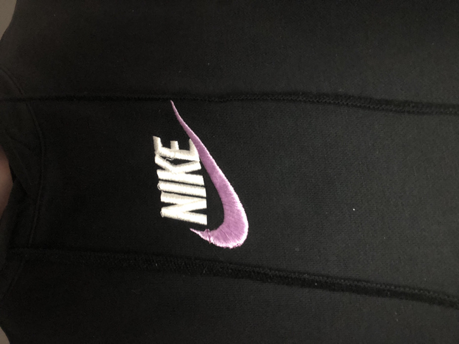 A close-up, slightly angled shot focuses on a black piece of clothing, possibly a hoodie, sweatshirt, or leggings, featuring an embroidered Nike logo. The garment is made of a soft, dark fabric, with a visible black stitched seam running horizontally across the middle, suggesting a panel design.
The embroidered Nike logo consists of the word "NIKE" rendered in white thread, and the iconic "swoosh" symbol positioned to the right of the text, embroidered in a light lavender or pastel purple thread. Upon closer inspection, some minor imperfections are visible in the embroidery. Specifically, there are a few loose threads or slight pulls in the white stitching on the letters 'K' and 'E' of "NIKE", and a small snag or unevenness at the top curve of the purple swoosh. The texture of the embroidery is slightly raised from the fabric surface.
No individuals or specific activities are depicted. The background is indistinct, indicating a close focus on the item. The lighting is soft and even, highlighting the textures and colors of the garment and embroidery. There are no distinct cues to time of day or weather within the image. While the image itself does not provide location cues, the context provided states the location is Moschato, Greece.