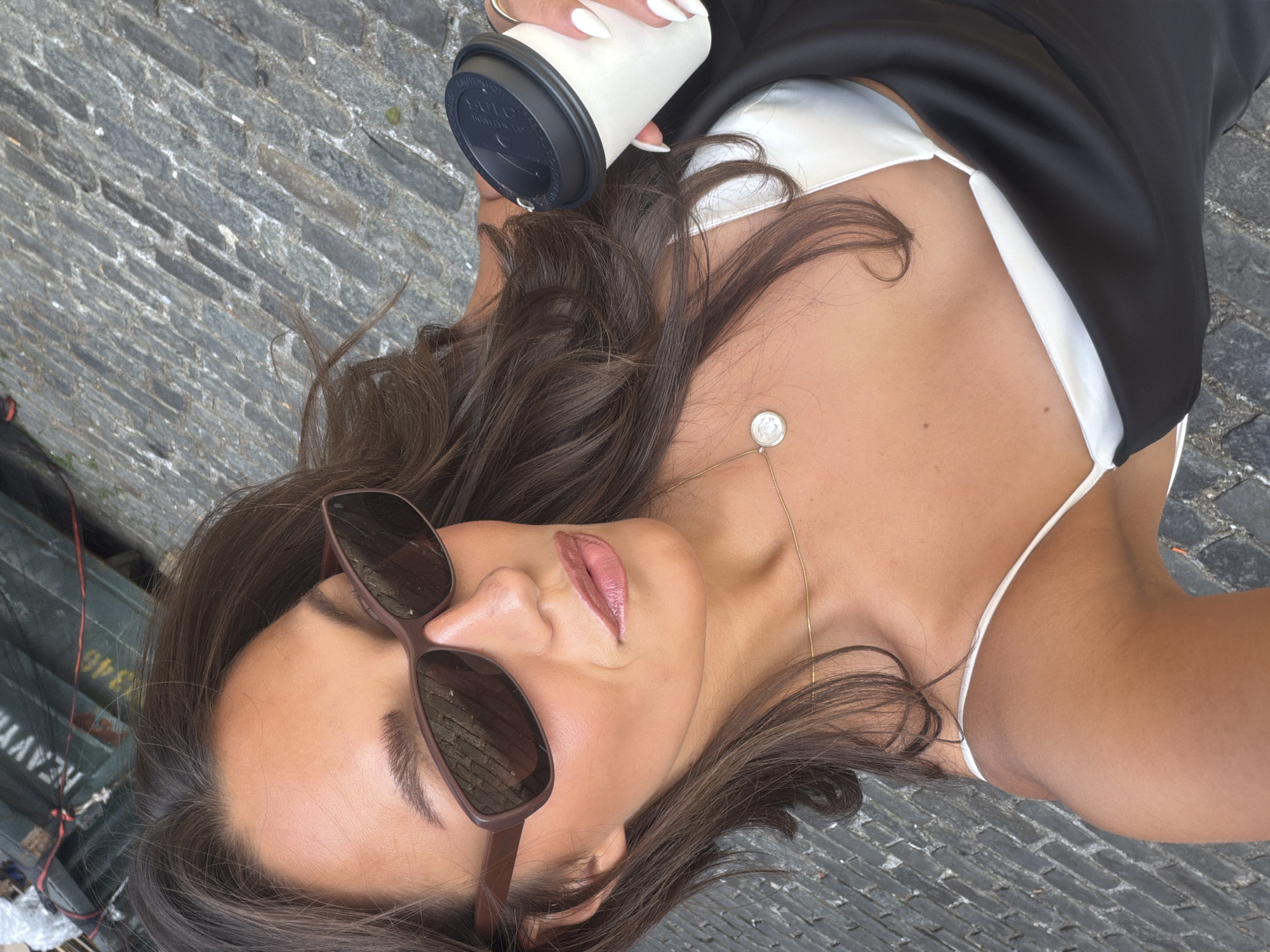 A woman with long, dark, wavy hair, wearing sunglasses and a black dress over a white camisole, holds a disposable coffee cup in her right hand. She is taking a selfie outdoors, with a cobblestone street and a green shipping container with the word "HEAVY" and the number "346" visible in the background. The scene suggests a casual outing in an urban environment.