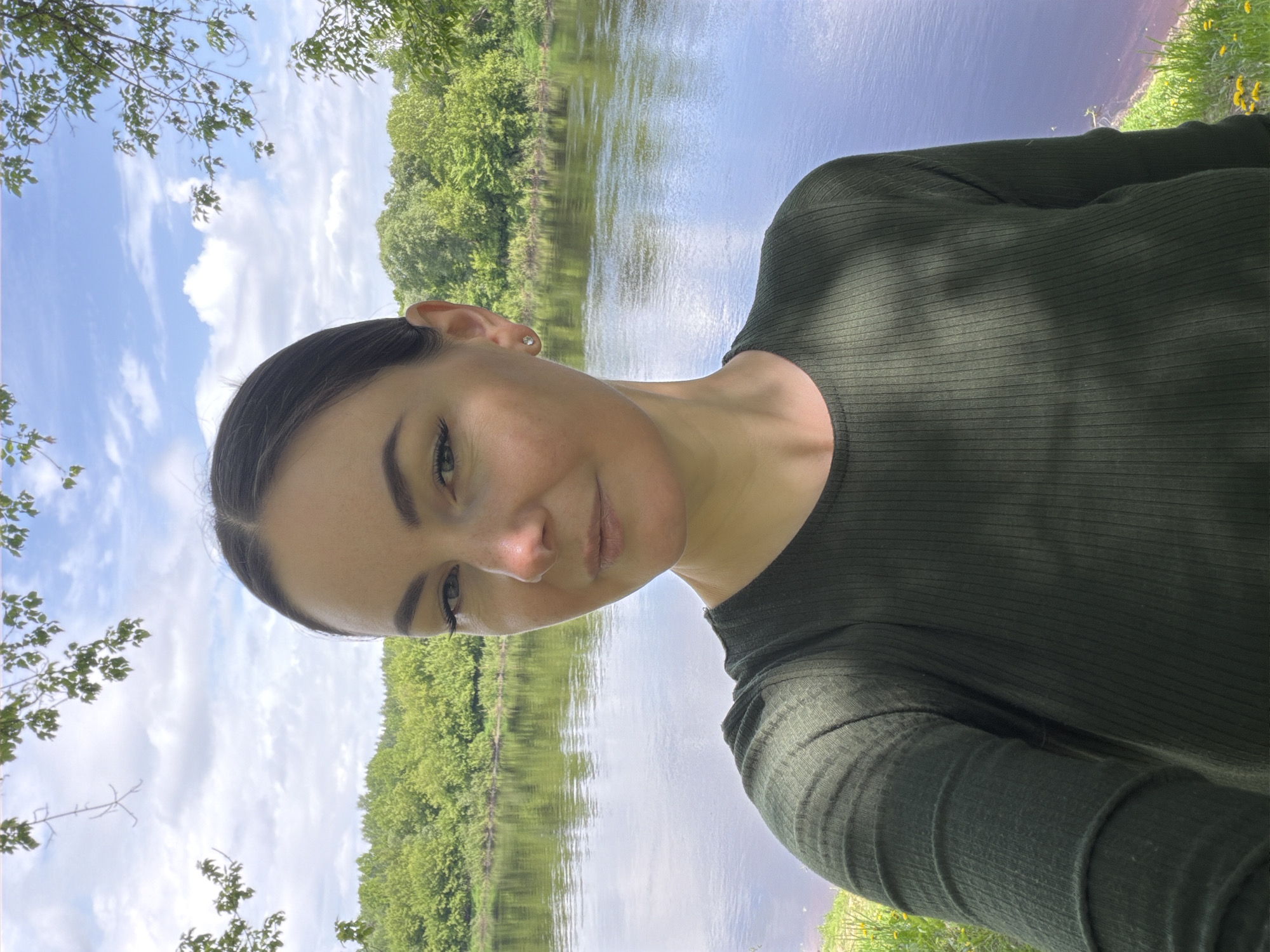 A woman is seen in a selfie-style photograph taken in an outdoor, riverside setting. She is dressed in a dark green, long-sleeved top and her face is angled towards the camera. Behind her, there is a river reflecting the sky and trees. The landscape features green trees lining the riverbank, with patches of blue sky and clouds visible overhead. There are also some yellow flowers in the foreground. The location of the photo is Unknown, Unknown.