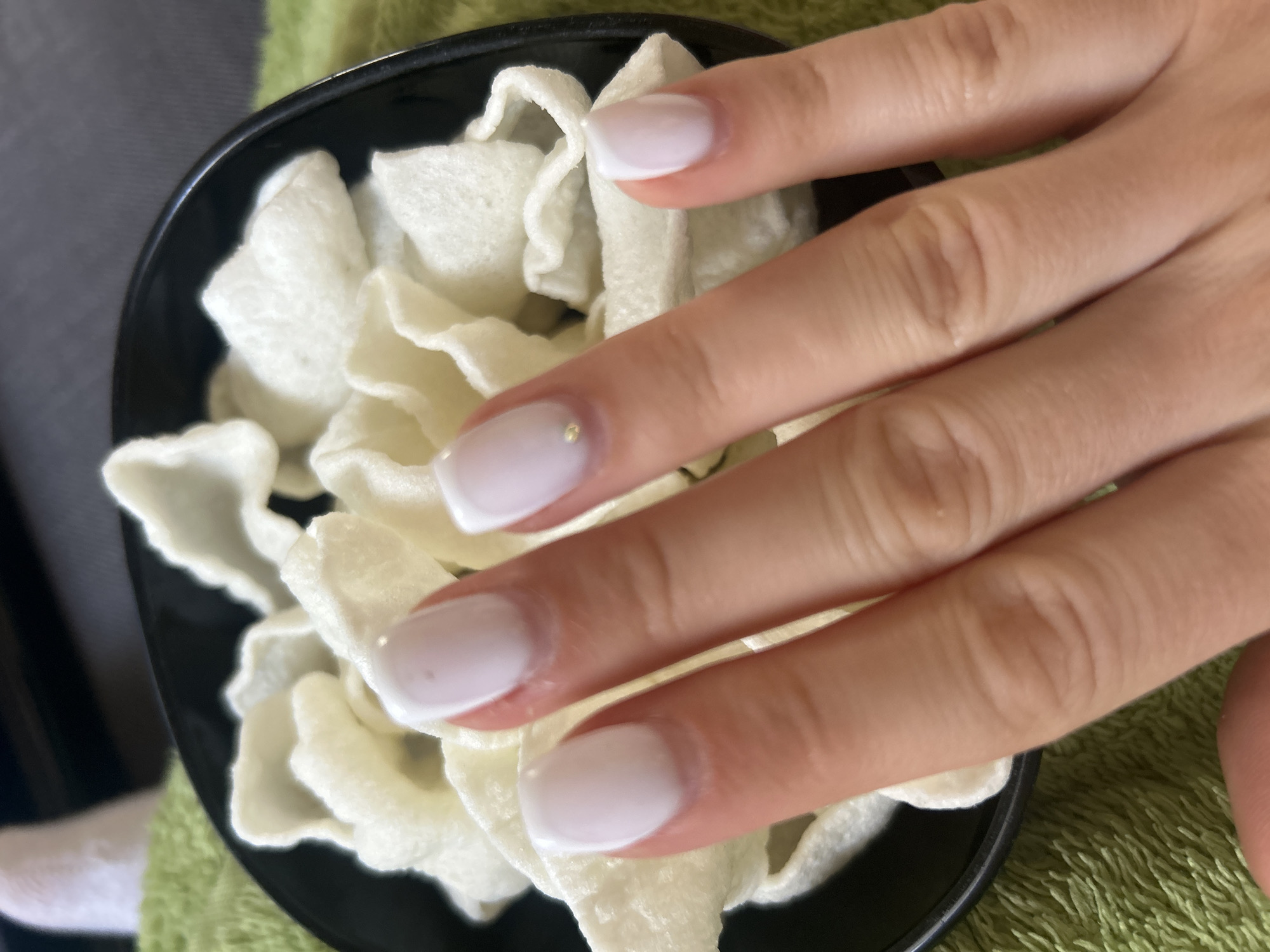 A close-up shot shows a hand with pale skin and nails with a white design, resting atop a black bowl filled with white, puffed snacks or chips. The background includes a green, textured surface, possibly a towel or fabric.