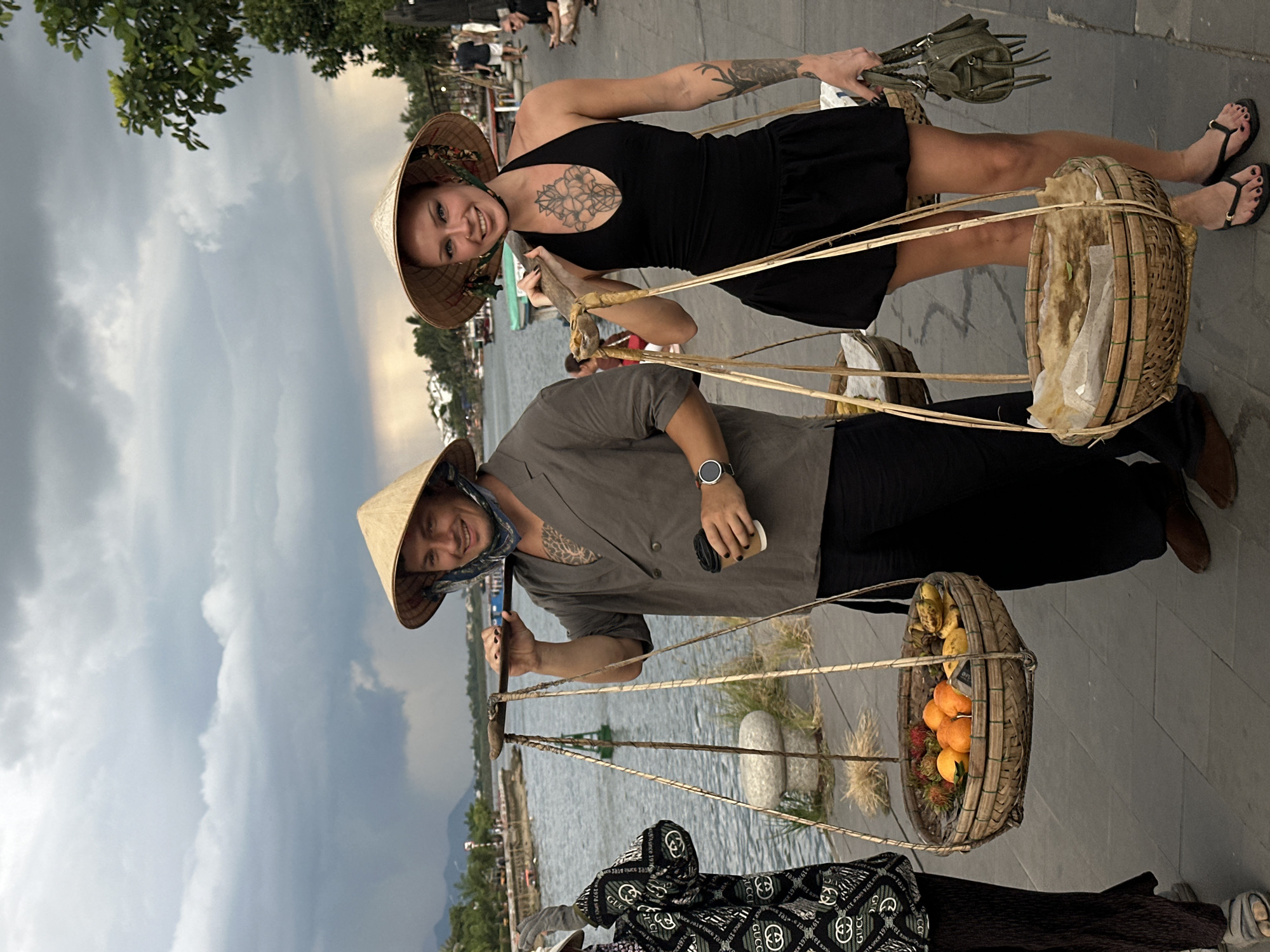 Two individuals, a man and a woman, stand on a paved walkway beside a river in Hoi An, Vietnam. Both wear conical hats, a traditional Vietnamese accessory. The man, on the left, wears a grey open-collared jacket over a dark garment and carries a yoke with two baskets filled with fruit, including rambutans, oranges, and bananas. He holds a disposable coffee cup in his right hand and has visible tattoos on his chest and arm. The woman, on the right, wears a black halter dress and also carries a yoke with baskets, one appearing to contain food items wrapped in paper. She has tattoos on her chest and arm and carries a small olive-green purse in her left hand. Behind them, the river flows under a cloudy sky, and buildings and greenery are visible on the opposite bank. In the background, other pedestrians are present. The scene suggests a bustling marketplace or riverside thoroughfare in the historic city.