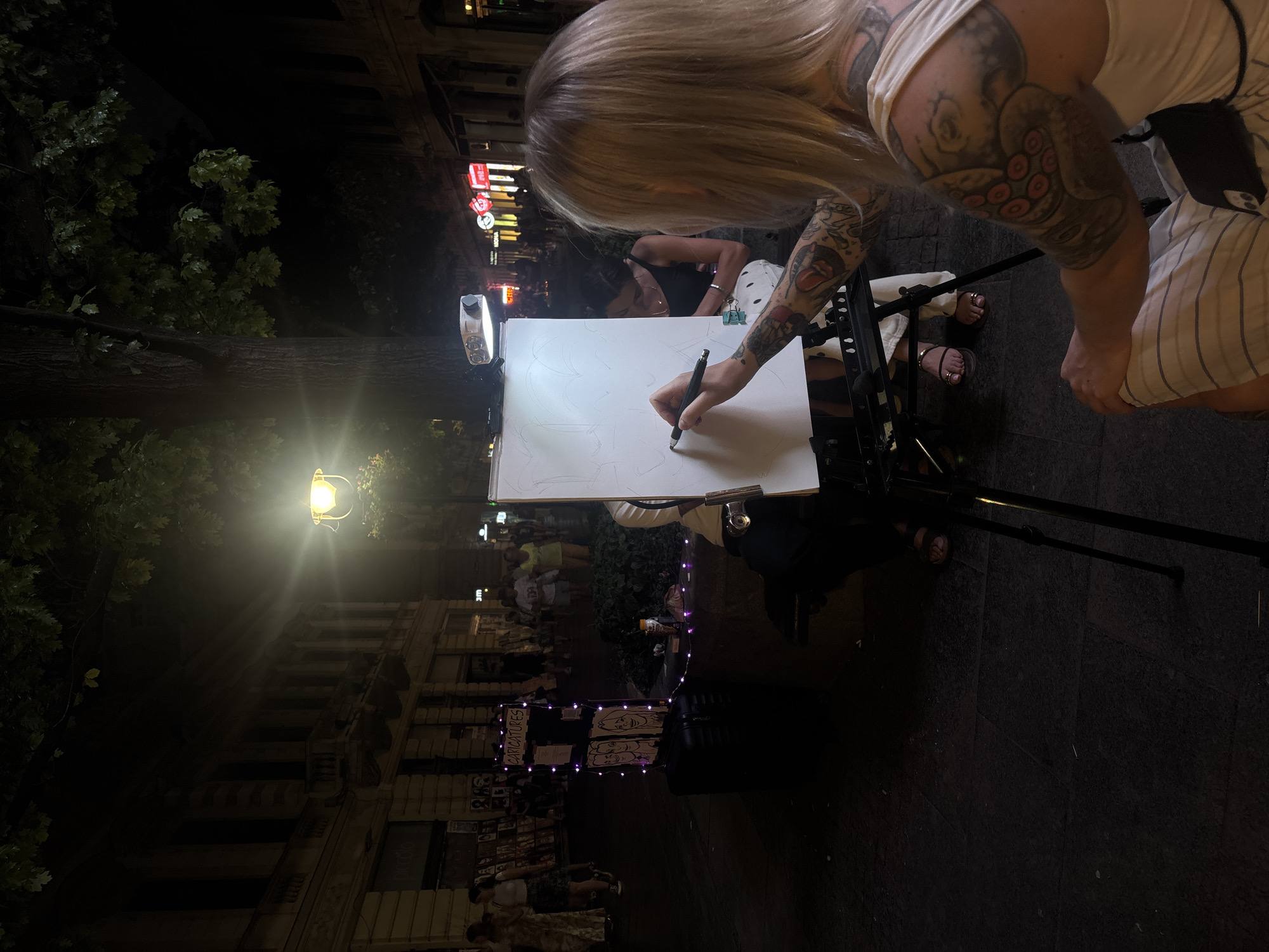 This is a photograph of an outdoor scene at night on a city street, likely in Belgrade, Serbia, given the architectural style of the buildings and the presence of a caricature artist. In the foreground, a person with blonde hair and extensive tattoos on their arms and back is seated, drawing on a large white canvas on an easel. They are holding a pen or pencil in their right hand and appear focused on their work. The canvas has a faint sketch of what seems to be a heart shape. The artist is wearing a white tank top and striped pants.
Behind the artist, another person in a black tank top is seated and partially visible, also appearing to be involved in the artistic setup. In the mid-ground, a stand displays framed caricatures and is decorated with purple fairy lights. This stand is positioned on a black suitcase. Further back, other people are walking along the street, illuminated by streetlights and neon signs from shops lining the buildings. The street is paved with dark stones. Trees with dark foliage hang over the scene from above, and a lamppost with a bright yellow light is visible on the right side of the frame. The overall atmosphere is that of a lively evening street scene where artists are showcasing their work.