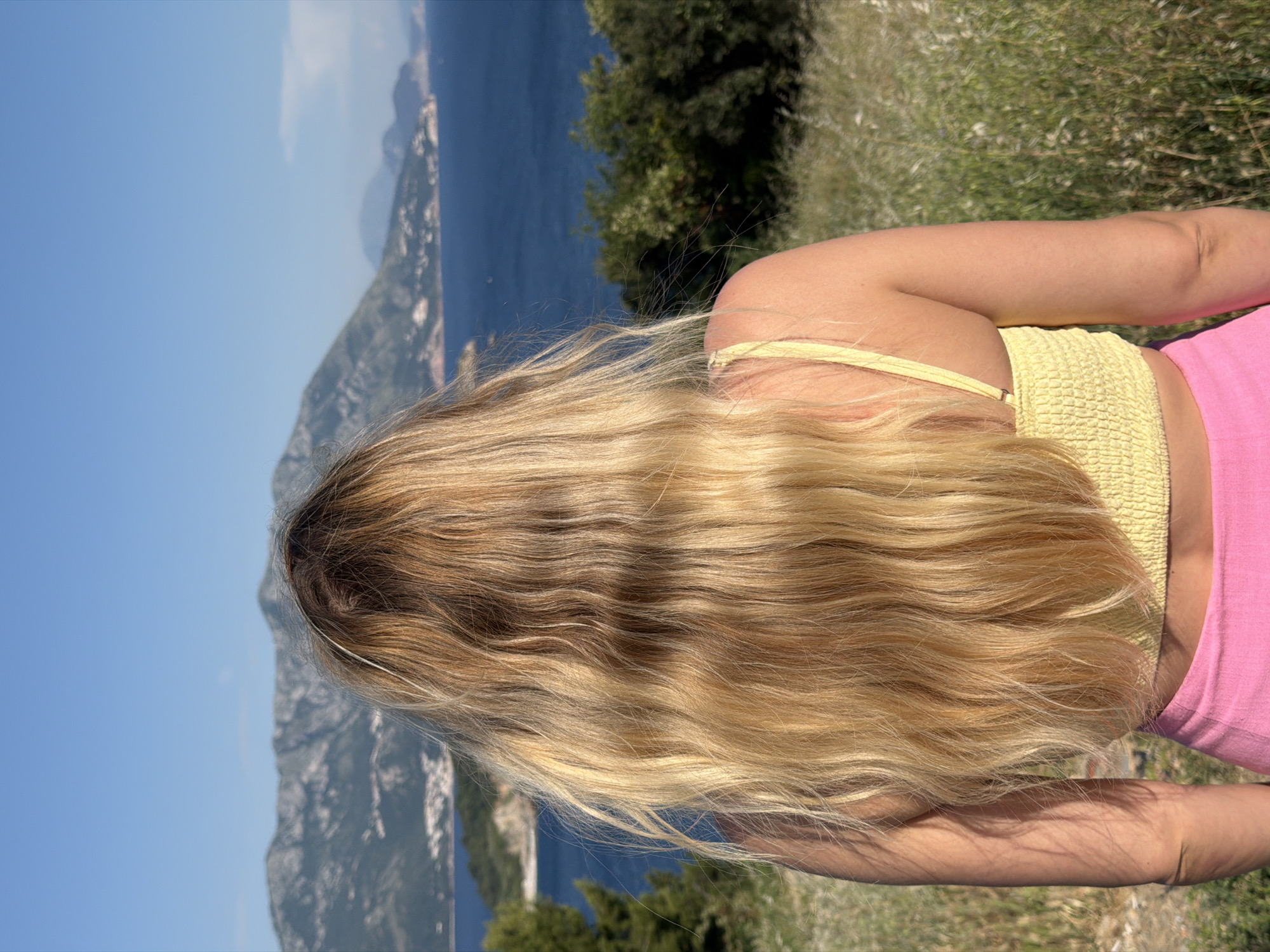 A medium shot depicts a woman from behind, showcasing her long, wavy blonde hair. She is wearing a yellow knit top with spaghetti straps and pink bottoms. The background reveals a scenic view, including a body of water, possibly the Adriatic Sea, and mountains, indicating an outdoor location. The grass and trees suggest the subject is in a natural setting with potential viewpoints overlooking Budva, Montenegro.