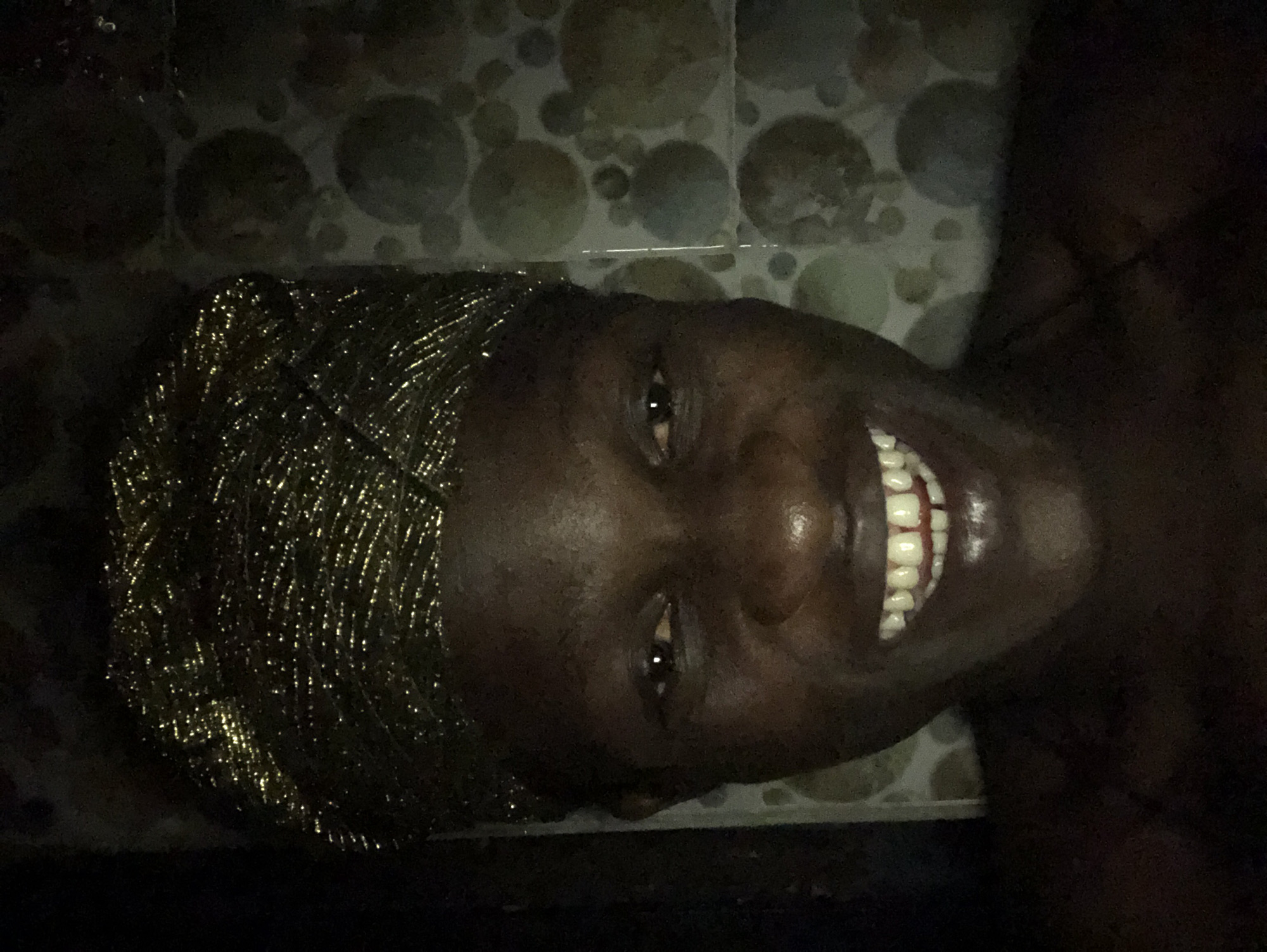 A close-up selfie of a dark-skinned person wearing a gold, textured headwrap is captured in a dimly lit indoor setting. The person is smiling, with their teeth visible. The background appears to be a tiled wall with a circular pattern. The location is identified as Ado, Nigeria.