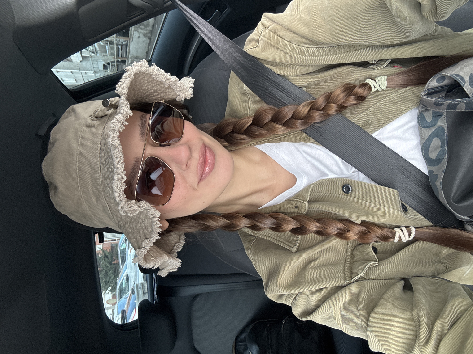 A woman is in a car, wearing a beige bucket hat, sunglasses, a white t-shirt, and a beige jacket. Her hair is braided, with white ties at the ends. She is wearing a seatbelt. The background appears to be an urban setting, visible through the car window, consistent with the city of Krasnoyarsk, Russia.