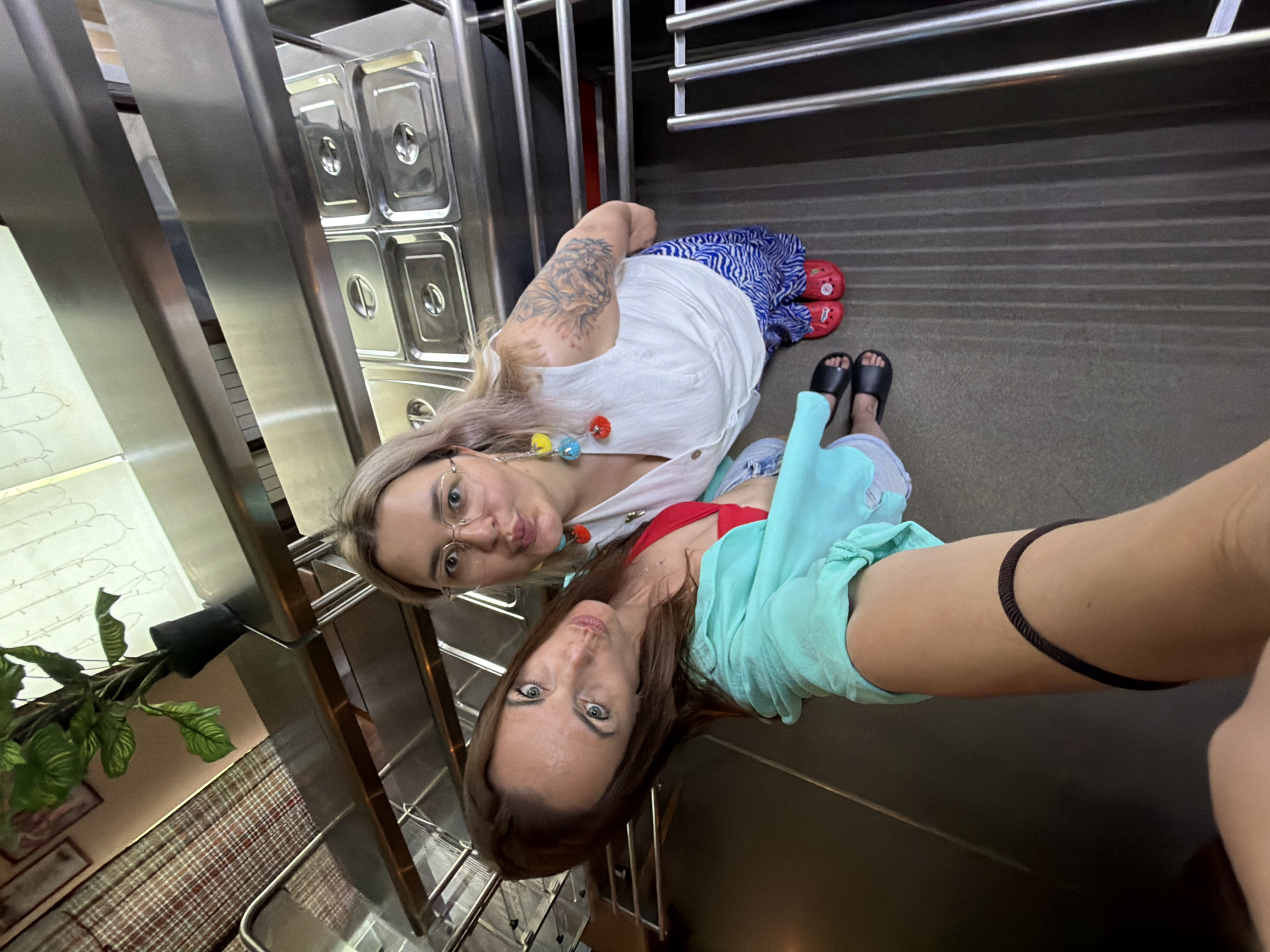 Two women are posing for a selfie in what appears to be a restaurant or buffet setting. The woman in the foreground is leaning towards the camera, looking sideways with her mouth pursed. She has brown hair, blue eyes, and is wearing a turquoise button-down shirt over a red bikini top and gray shorts. A black cord is wrapped around her left arm. The woman behind her is looking directly at the camera with her mouth puckered, wearing glasses and a white sleeveless top. She has blonde hair and is wearing blue patterned pants and red Crocs. She has a tattoo on her left arm and colorful earrings. Behind them, stainless steel serving trays are visible on shelves, along with a potted plant. The floor is dark and ribbed. There is no indication of Birobidzhan, Russia in the image.