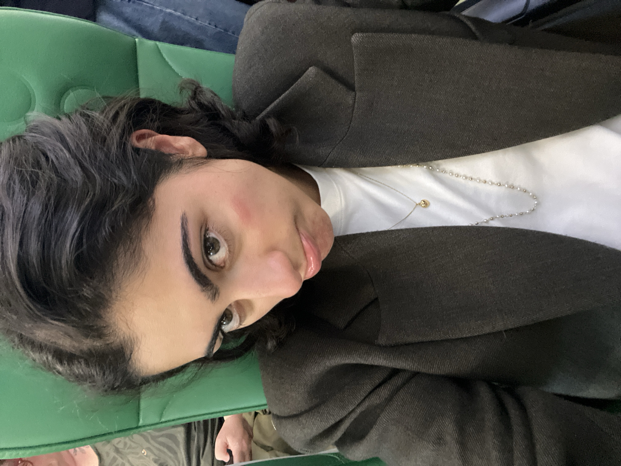 A person is lying on a green cushion with a jacket over a white shirt, wearing two necklaces. The location is not identifiable.