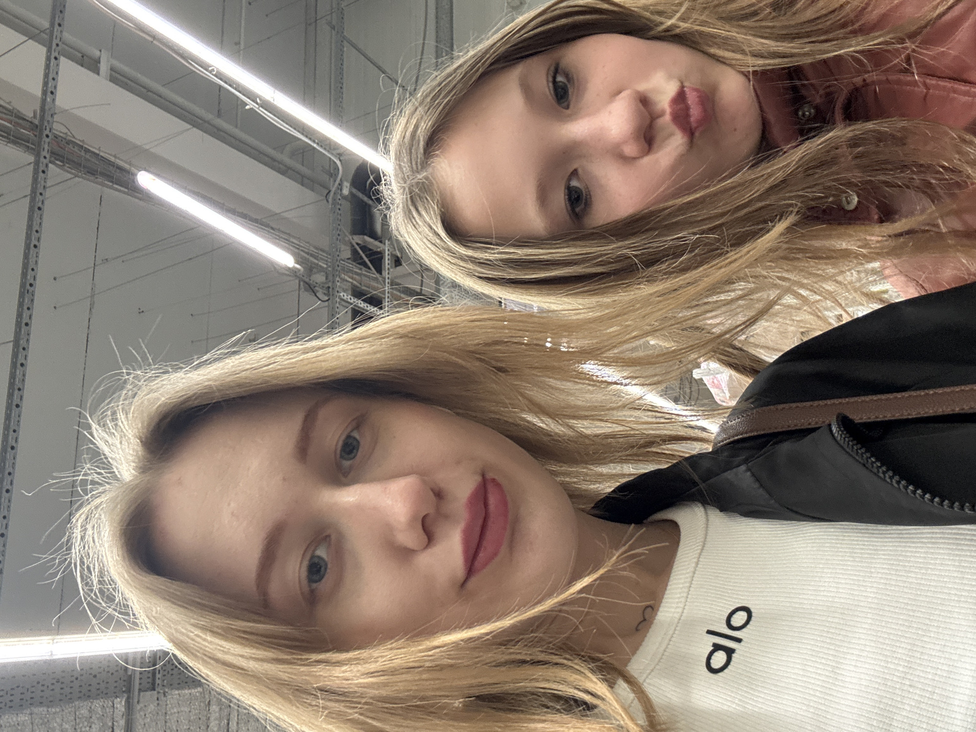 A close-up image depicts two young females posing for a photograph in what appears to be an indoor commercial or retail establishment in Rostov-on-Don, Russia.
The female on the left, positioned closer to the camera, has long, straight blonde hair and blue eyes. She wears a white ribbed long-sleeved top with the word "alo" printed in black on the chest. A small, dark, heart-shaped tattoo is visible on her neck below her left ear. She also wears a black jacket, and a brown bag strap is visible over her right shoulder. Her expression is neutral, and her lips are tinted with a rose-colored product.
The female on the right, positioned slightly behind and to the right, has long, straight light brown hair and blue eyes. She wears a reddish-brown or dusty rose-colored jacket. Her lips are pursed in a "kissing" pose.
The background consists of a light-colored, possibly white or light grey, ceiling with exposed industrial infrastructure. This includes metallic cable trays, conduits, and multiple bright, linear fluorescent light fixtures, indicating an overhead utility grid typical of large commercial or industrial spaces. The lighting is uniform and bright, illuminating the subjects from above.