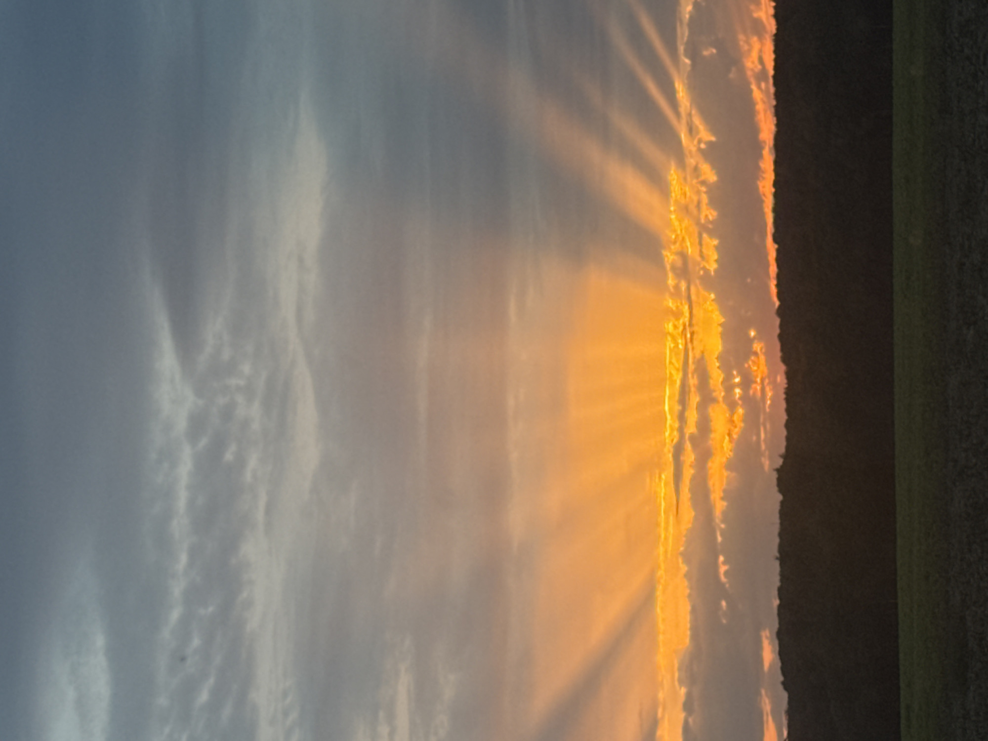 The image shows a sunset over a rural landscape. The sky is filled with dramatic clouds illuminated by the setting sun, which is partially obscured by the clouds, casting rays of light downwards. The foreground features a dark silhouette of a tree line or forest, and below that, a cultivated field. The scene appears to be in a natural or agricultural setting, not a densely populated urban area. The image does not depict any people or specific structures that would identify it as being within the city of Eyendorf, Germany.