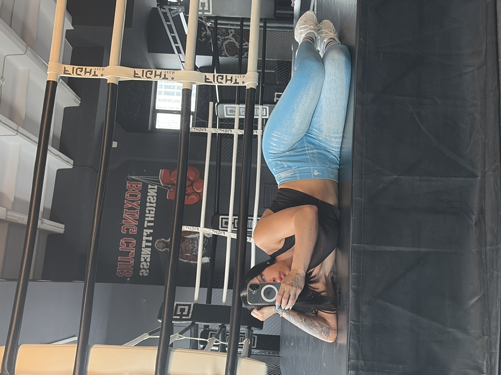 A woman is lying on her back on a gym floor, with her legs bent and propped up against what appears to be a metal structure. She is taking a selfie with her phone, which is held up to her face. She is wearing a black sports bra and light blue leggings with a textured pattern. Her hair is dark and long, and she has tattoos on her arm.
The setting is a boxing club, indicated by the sign on the wall that reads "BOXING CLUB" and "INSIGHT FITNESS". There are also boxing gloves visible on the wall behind her, and metal poles and netting are part of the gym equipment. The overall impression is of a workout session in a modern fitness facility. The lighting suggests it could be daytime. The woman's pose and selfie indicate a sense of self-awareness and perhaps a moment of rest or reflection during her workout.
