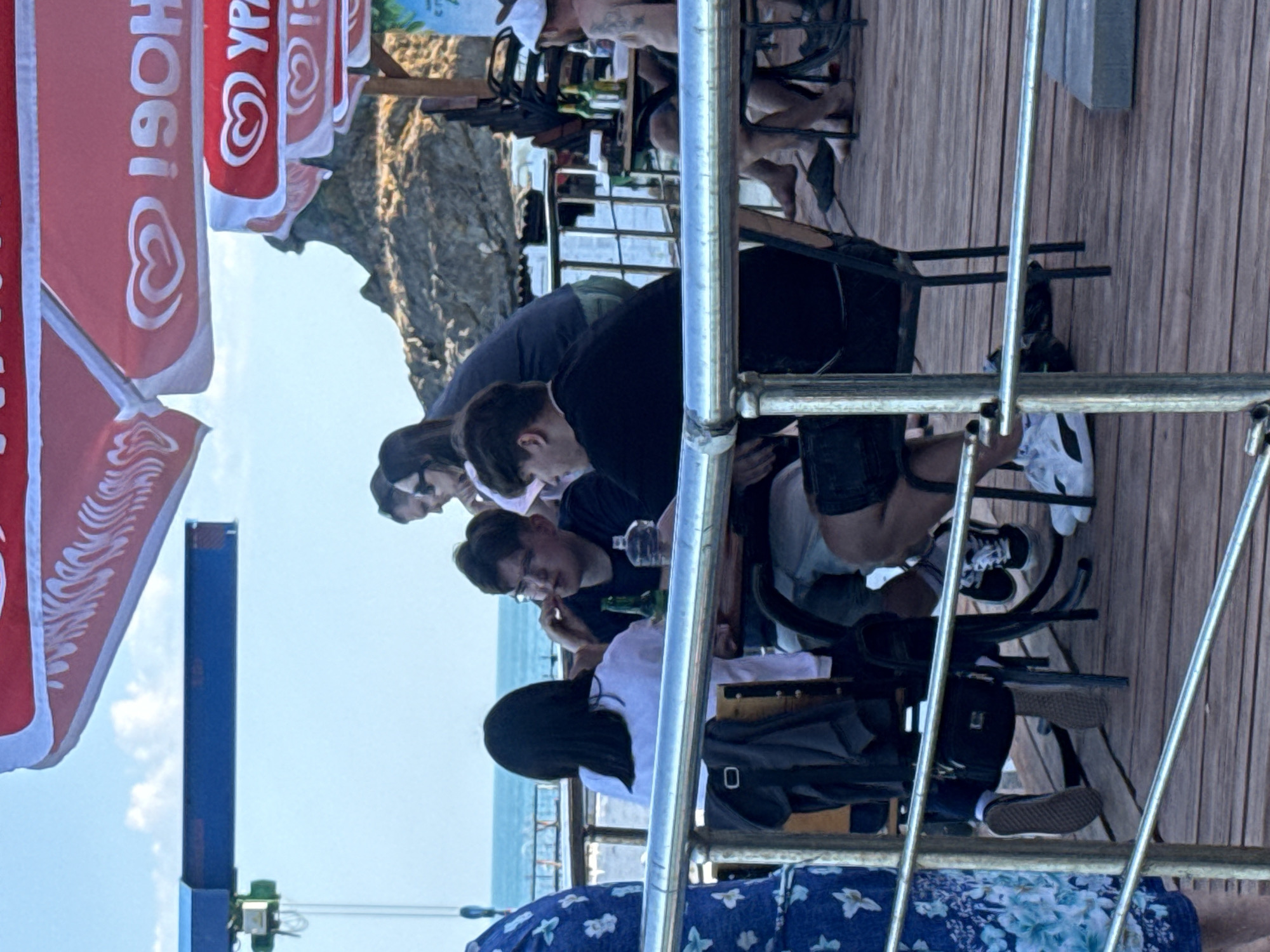 The image shows a group of people sitting at tables on a wooden deck. The scene appears to be outdoors, possibly at a seaside cafe or restaurant, indicated by the ocean in the background and large red and white umbrellas providing shade. In the foreground, a metal railing separates the viewer from the deck.
On the deck, several individuals are seated. Two young men are at one table, with one wearing glasses and holding what appears to be a cigarette. A bottle and a glass of water are on the table. Another young woman with dark hair is seated at the same table. Further back, a woman in a blue shirt is standing. To the right, another table has several people seated, including a man with a white cap and tattoos on his arm. Bottles are also visible on this table. The ground beneath the tables is made of wooden planks.
In the background, a rocky incline is visible, and the sky is a light blue with some clouds. There is also some scaffolding or construction element on the left side of the image. The overall impression is a casual gathering of people enjoying themselves in an outdoor setting.