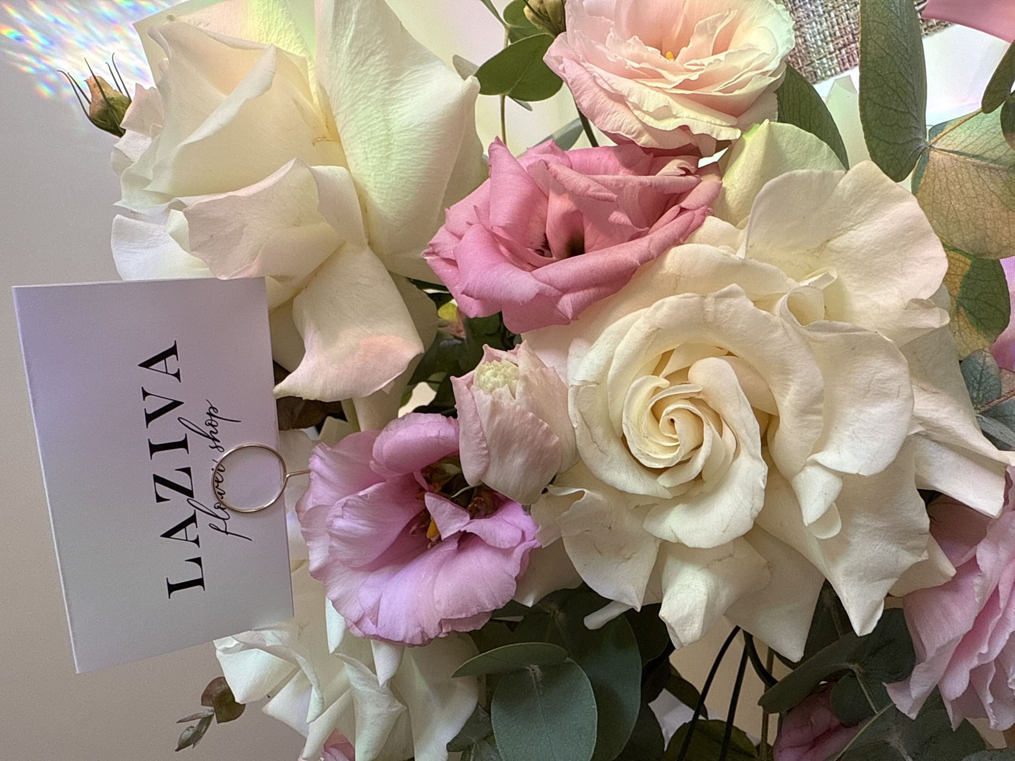 An overhead shot captures a bouquet of cream and pink roses, interspersed with eucalyptus leaves. A small white card, clipped to the arrangement with a gold wire holder, displays the text "LAZIVA flower shop" in black serif font and a script font, respectively. The flowers are artfully arranged, with some petals slightly unfurling, revealing their delicate textures. The background is a soft, neutral tone, with a subtle prism effect creating streaks of rainbow light on the right side of the frame. The overall impression is one of elegance and natural beauty.