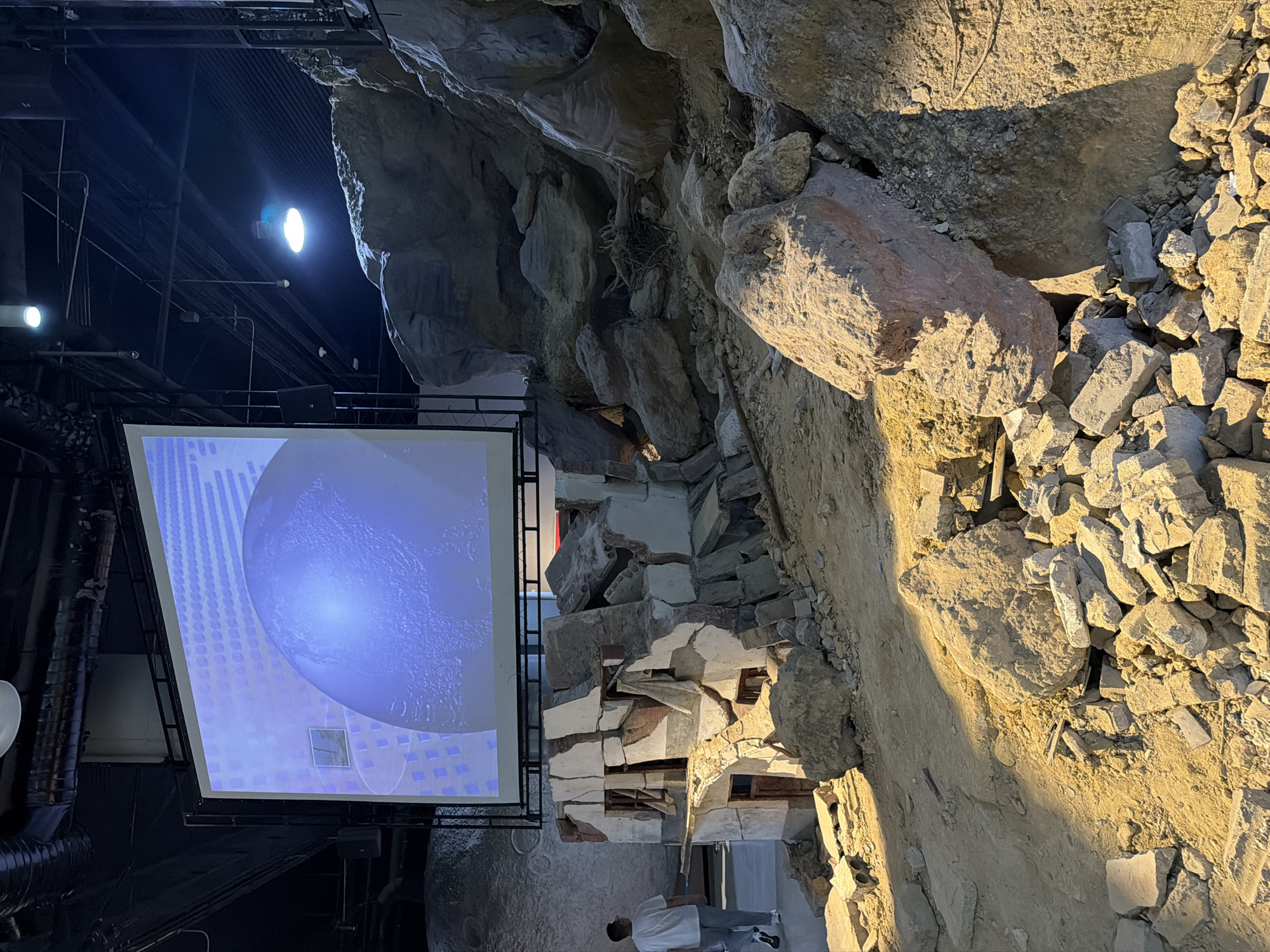 The image depicts an indoor scene with a large projection screen displaying an image of a blue sphere, possibly Earth. In the foreground, there is a simulated scene of a partially demolished structure made of concrete and bricks, with large rocks and debris scattered around it, suggesting a post-disaster or construction site environment. A person in a white t-shirt and grey pants is walking away from the camera towards the left side of the frame, past the demolished structure. The environment appears to be a public space, possibly an exhibition or museum, given the projection screen and the staged debris. The lighting is artificial, with spotlights illuminating the scene. The location, 江北城街道, China, suggests an urban setting.