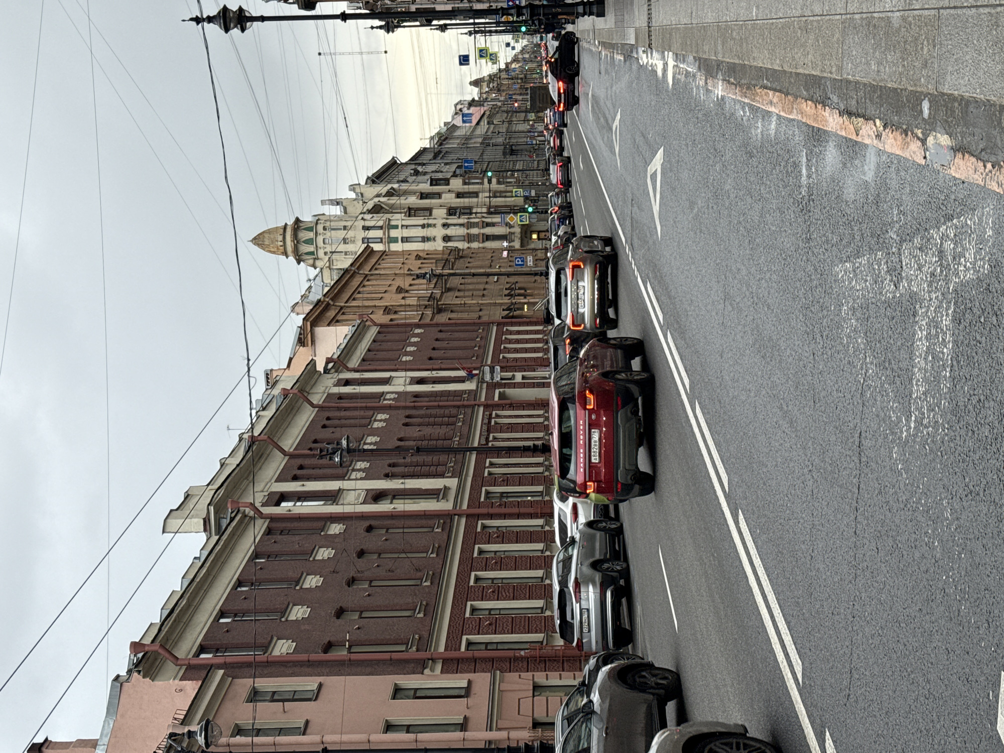 The image depicts a street scene in Saint Petersburg, Russia. The view is along a multi-lane road with cars lined up, suggesting traffic congestion. The buildings lining the street are a mix of architectural styles, with some featuring ornate facades and classical details. The sky is overcast, providing a muted light to the scene. Overhead electrical wires span the street. A golden dome is visible in the background, atop one of the buildings.