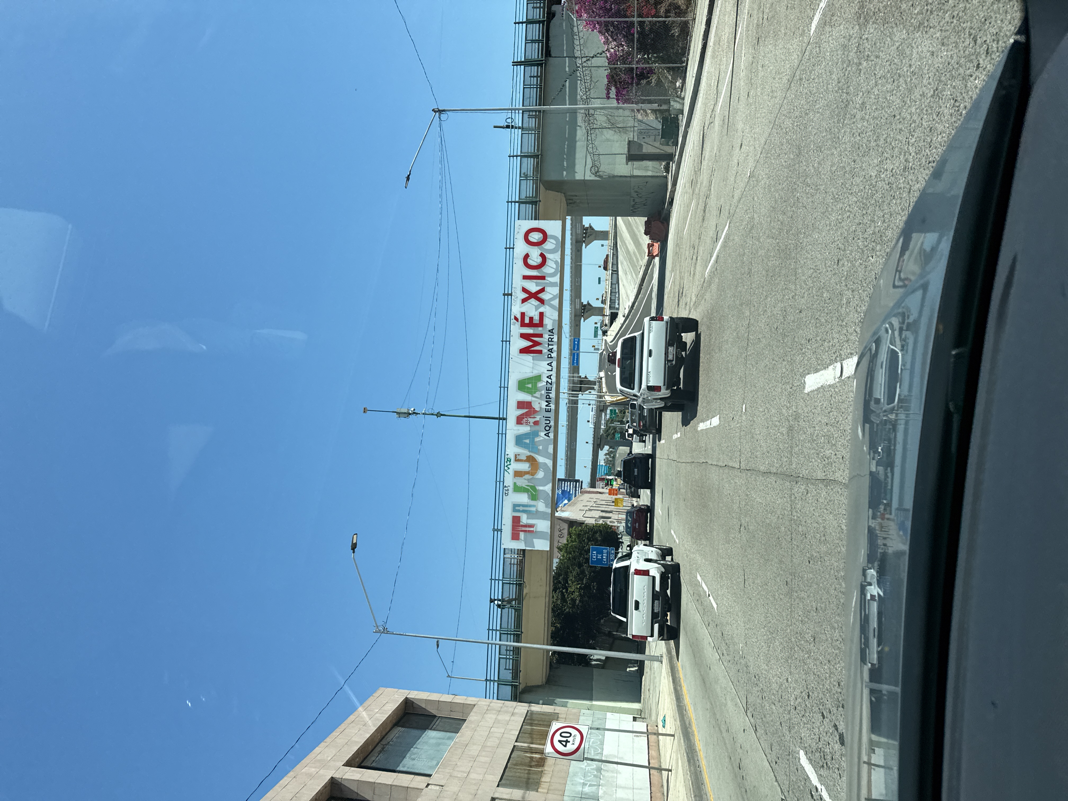 The image depicts a street in Tijuana, Mexico, specifically under a bridge that welcomes visitors to the city. The sign above reads "TIJUANA MEXICO" in large, colorful letters. Below this, in smaller text, it says "AQUI EMPIEZA LA PATRIA."
The view is from inside a vehicle, looking out through the windshield. In front of the vehicle, there are several other cars and trucks traveling in the same direction. The road is paved and marked with white lines.
To the left of the road, there is a building and a street sign indicating a speed limit of 40. To the right, there is a fence and some greenery.
The sky is clear and blue, suggesting it is daytime. The overall impression is of a busy street scene in a border city.