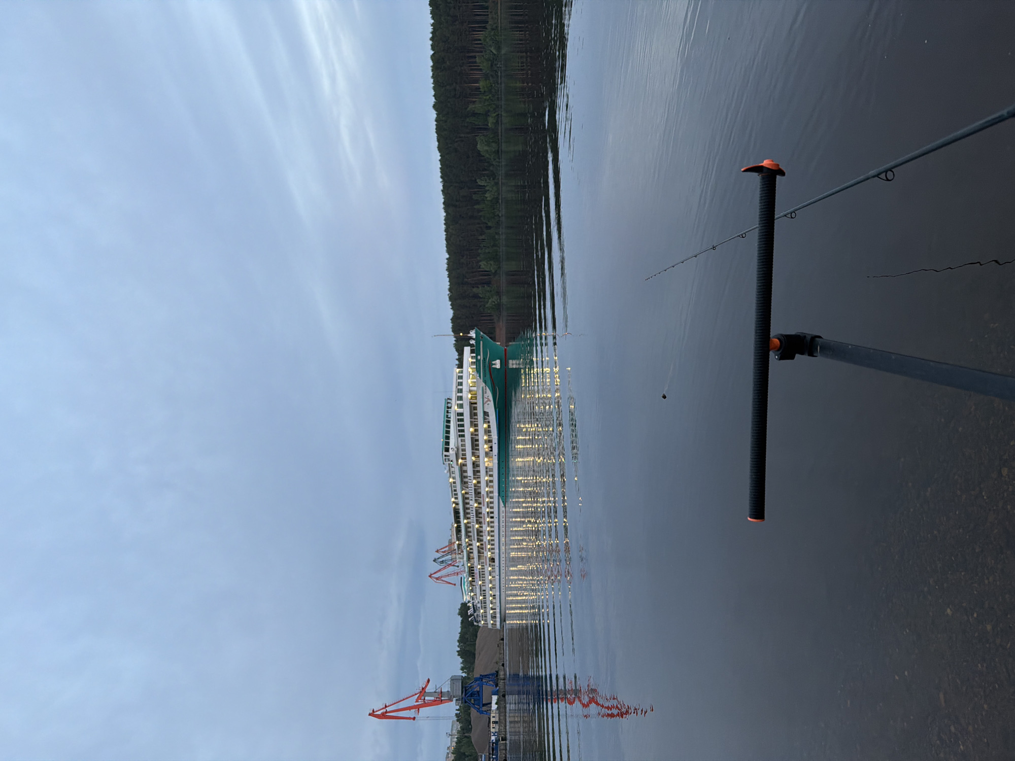 The image depicts a body of water, possibly a river or large lake, with a forest visible on the opposite shore. A large passenger ship illuminated with lights is docked near the shore. Cranes are positioned nearby. In the foreground, there is a fishing rod held by a rod holder. The image was taken in the city of Chaikovsky, Russia.