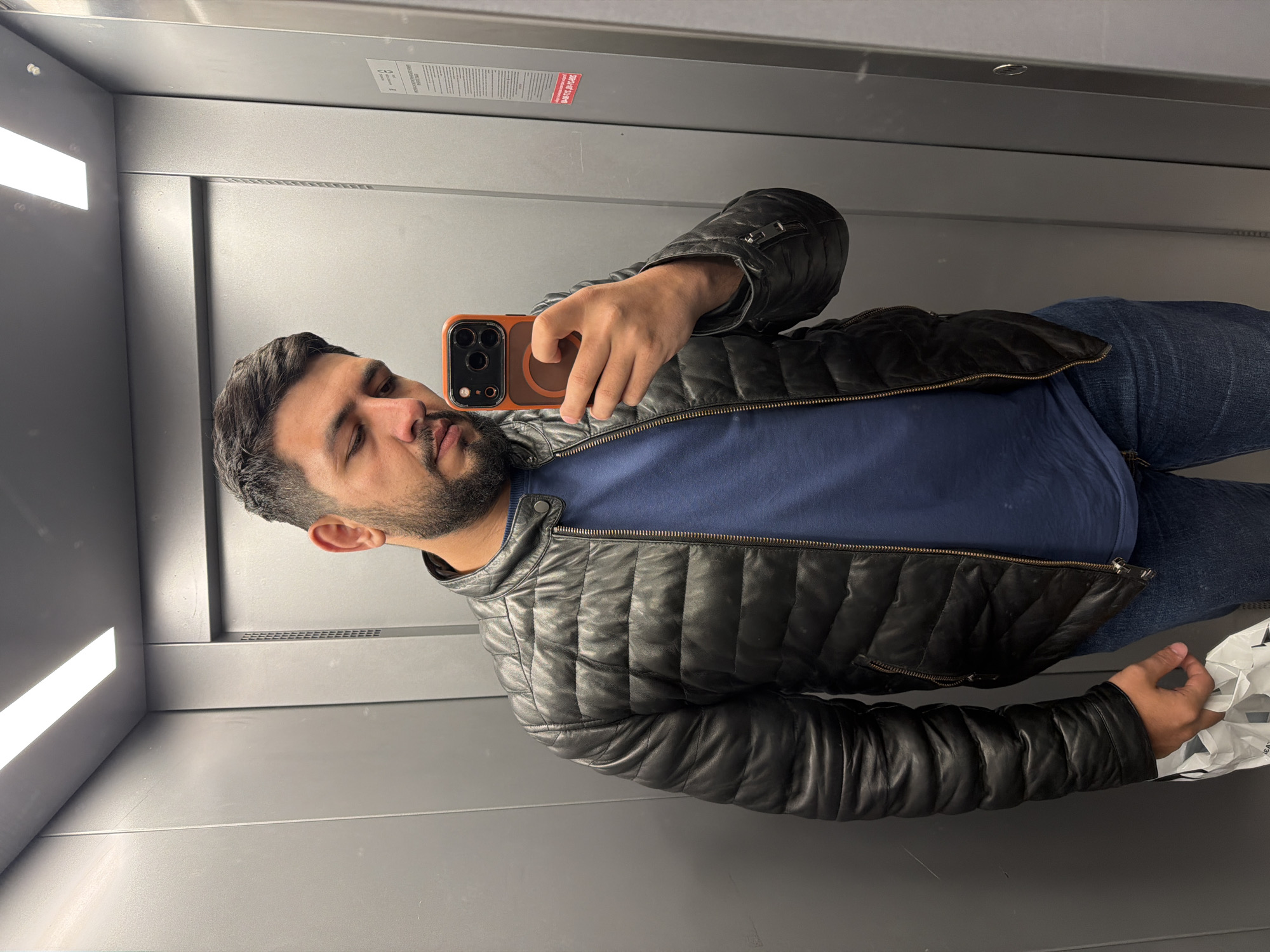 A man is taking a selfie in an elevator. He is wearing a black leather jacket over a blue shirt and blue jeans. He is holding an iPhone in his right hand, with an orange phone case. The elevator has gray walls and a rectangular light fixture on the ceiling. The elevator's interior appears generic, with no specific details indicating a location in Krasnoyarsk, Russia.