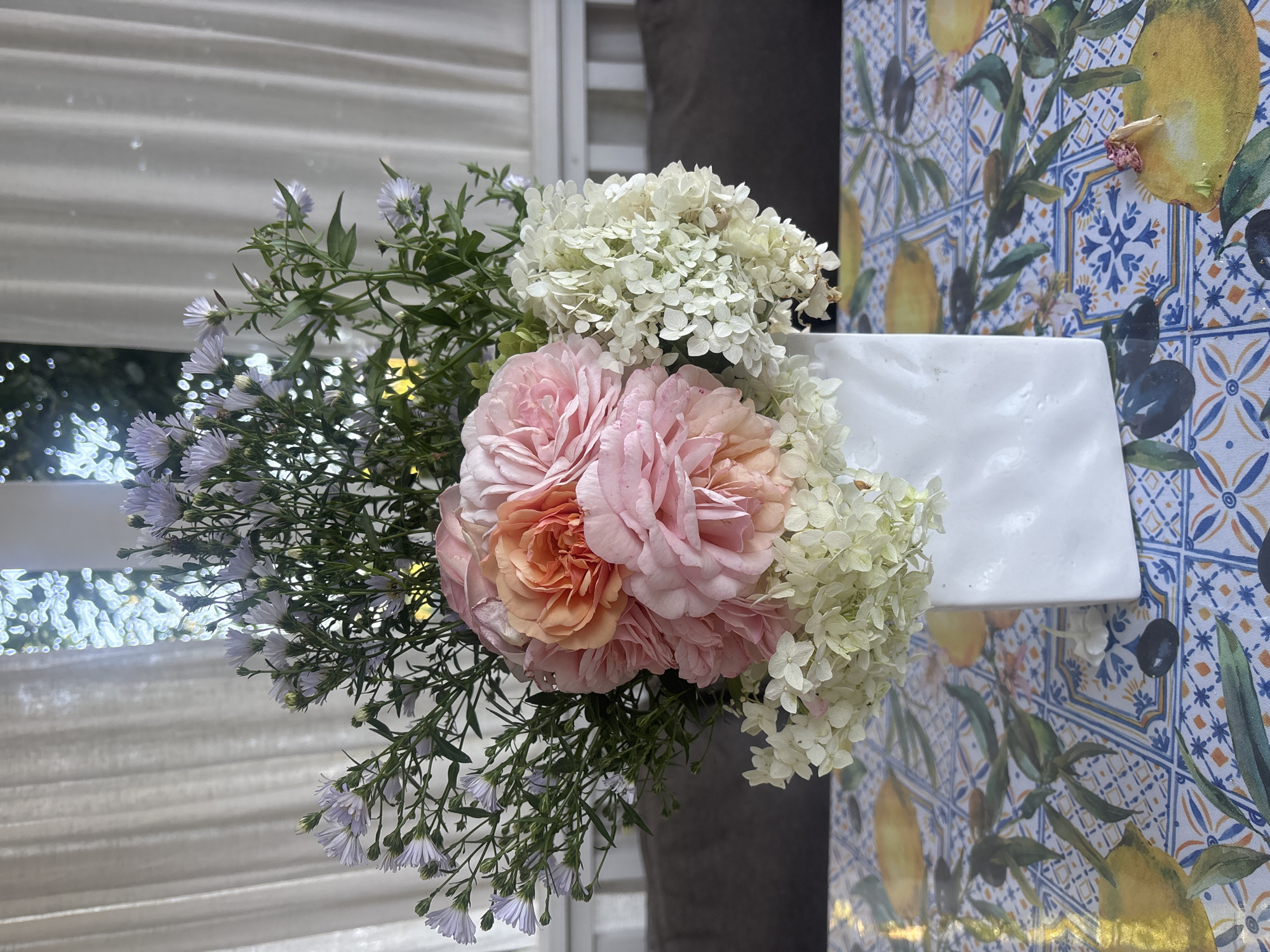 A vase of flowers is displayed on a table in what appears to be a sunroom or dining area. The vase is white and square with a textured surface, and it contains a bouquet of pink and peach-colored roses, small white flowers, and delicate purple aster-like flowers. The bouquet is lush and full, with greenery interspersed throughout. The table is covered with a tablecloth that features a colorful pattern of lemons, olives, and blue and white tiles. In the background, sheer white curtains are drawn, and sunlight streams through them, creating a soft, diffused light. Behind the curtains, hints of greenery and trees are visible, suggesting an outdoor setting. The overall impression is one of a bright, airy, and elegantly decorated space. The specific location of Лотос Снт (марьино), Russia is not discernible from the image itself.