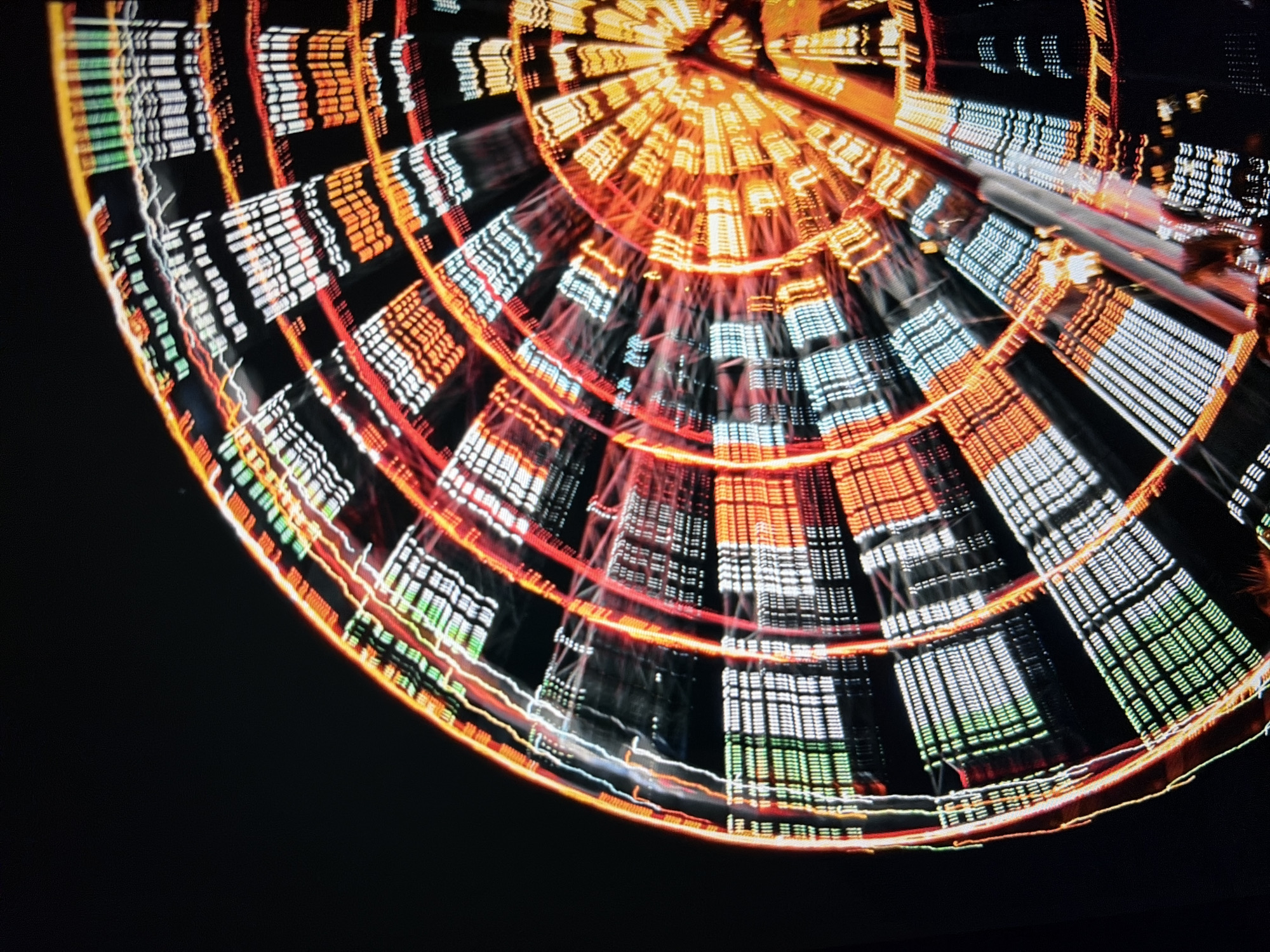 This is a long-exposure, close-up, abstract shot of a Ferris wheel at night. The image is dominated by concentric circles of colorful lights from the wheel, creating a swirling, motion-blurred effect. The lights are primarily red, orange, white, and green, with streaks and trails of color radiating outwards from the center. The background is completely black, emphasizing the illuminated structure of the Ferris wheel. The image appears to be taken from the ground, looking up at the massive structure. There are no visible people or identifiable landmarks, as the focus is entirely on the abstract patterns created by the light and motion.