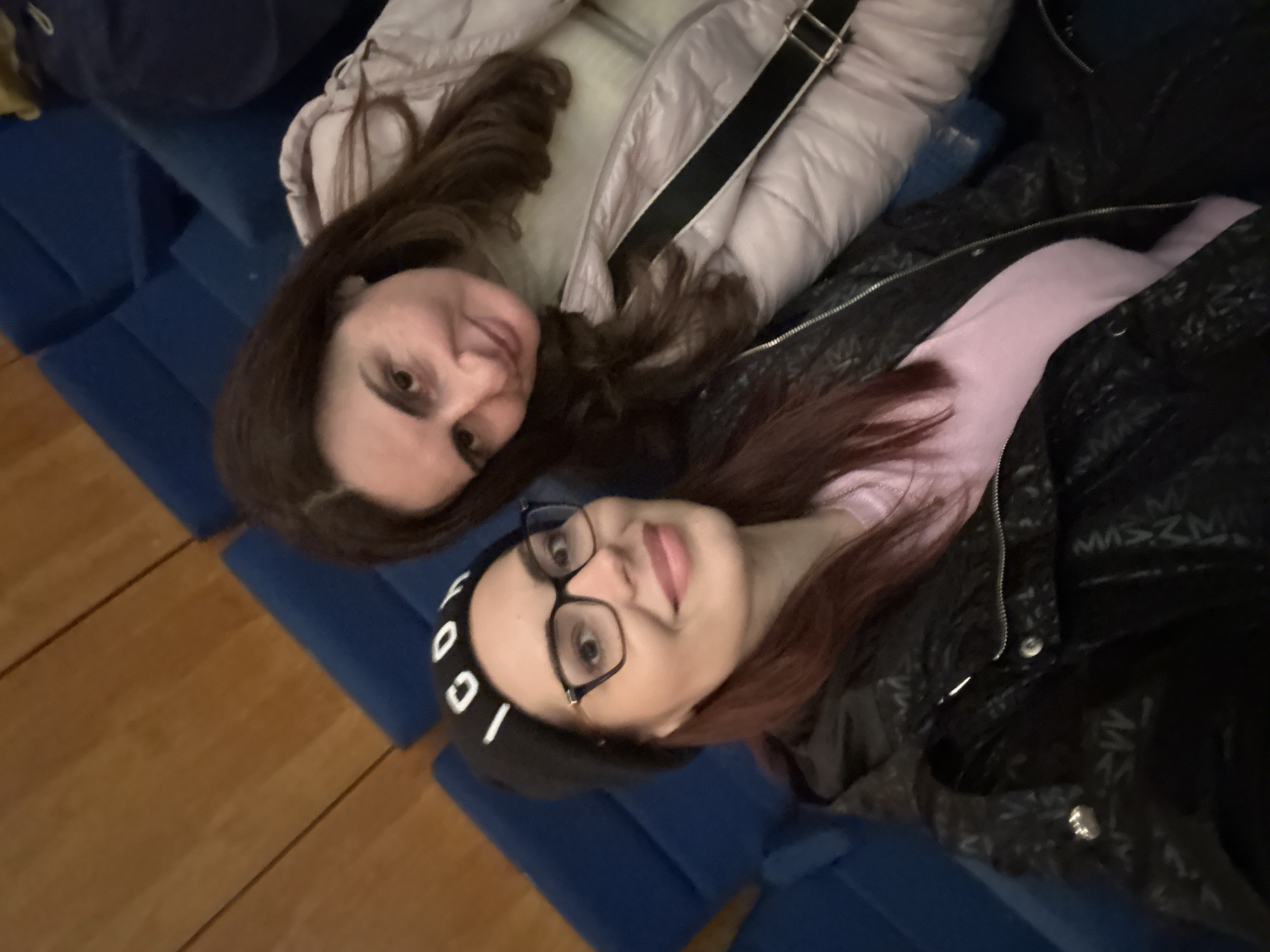 Two women lie on blue, rectangular cushions. One woman has long, brown hair and is wearing a light-colored, puffy jacket with a black strap. The other woman has long, reddish-brown hair, glasses, and a black beanie with white lettering. She is wearing a light purple shirt and a black jacket with silver zippers and buttons. The cushions are placed on a wooden floor. The location is in Montana, Bulgaria.