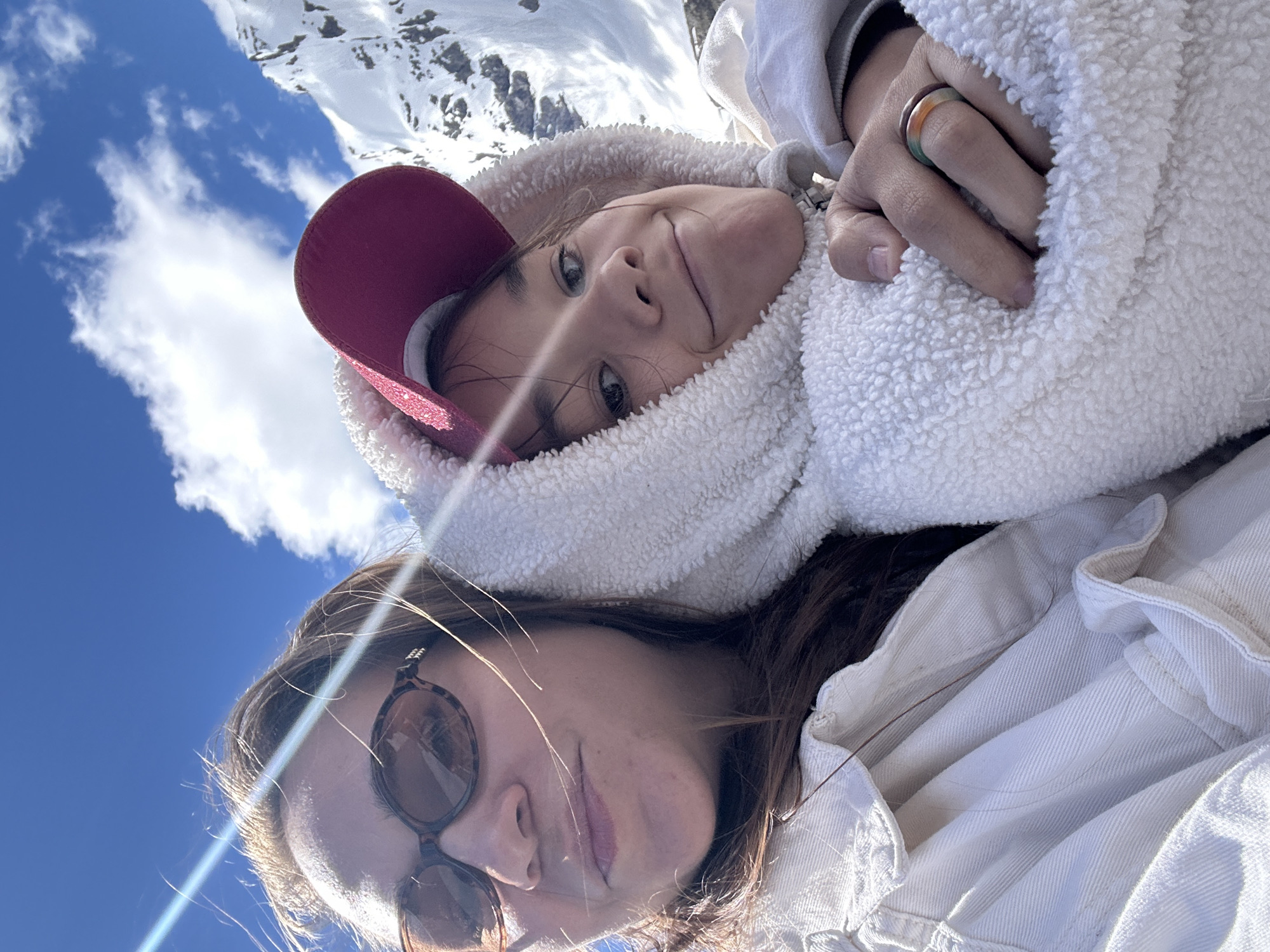 Two people, one appearing older and wearing sunglasses and a light-colored jacket, and a younger person wearing a red baseball cap and a white hooded fuzzy jacket, are taking a selfie outdoors. They are embracing, with the younger person's arm wrapped around the older person. The younger person is wearing a ring with multiple colored bands on their left ring finger. In the background, there are snow-capped mountains under a bright blue sky with white clouds. The sun is shining, creating lens flare across the image. The location is the city of Terskol, Russia.