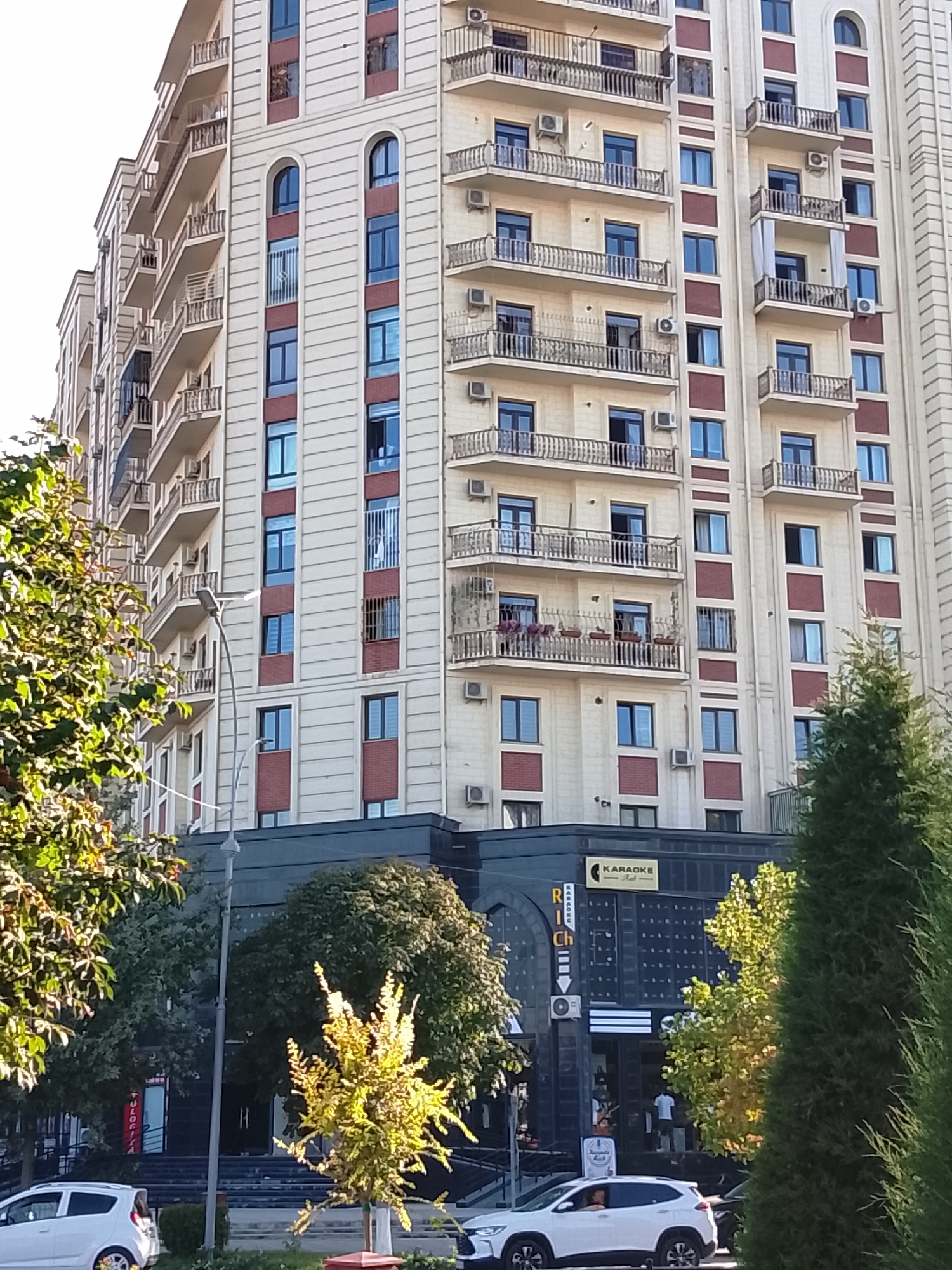 The image shows a multi-story building in Fergana City, Uzbekistan. The building has a light-colored facade with dark red accent panels and balconies with ornate railings. Windows are visible on each level. In front of the building, there are trees with green and yellow foliage. A street light stands near the left side of the image. At the base of the building, there is a dark-colored commercial establishment with a sign that reads "KARAOKE". A white car is parked on the street in front of the establishment. The overall scene depicts an urban environment with a focus on the architecture of the building and its surroundings.