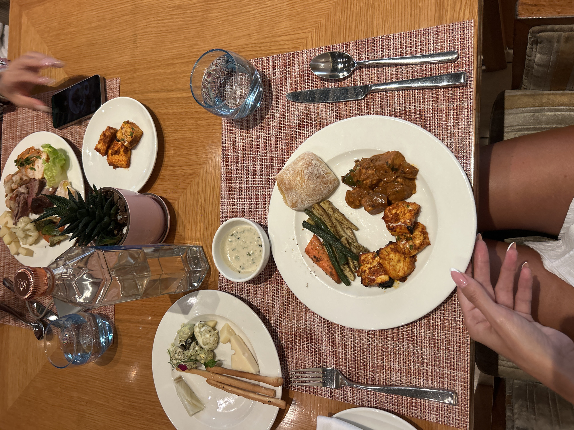 An overhead medium shot captures a dinner spread on a wooden table. In the foreground, a woman's hands with manicured nails rest on her lap, partially obscured by a white napkin. The table is set with two white plates of food, a clear bottle of liquid, a small potted plant, and glassware.
The plate closest to the foreground features a selection of Indian cuisine, including cubed paneer tikka, a rich, brown curry, and grilled vegetables like carrots and green beans, alongside a bread roll. A fork, knife, and spoon are placed to the right of this plate on a red and white patterned placemat.
Further back on the table, another white plate displays breadsticks, cheese, and a dip with green herbs. Adjacent to this is a small white bowl of a creamy sauce. A glass of water sits to the left of the bottle. The bottle, shaped like a faceted diamond, contains a clear liquid and has a metallic rose-gold cap. A small green succulent plant in a pink pot is placed between the bottle and a smartphone.
A third plate, positioned further away, holds a variety of appetizers, including what appears to be sliced meat, chicken, shrimp, and lettuce. A hand with blurred fingers is reaching towards this plate, suggesting someone is in the process of selecting food. Two glasses of water are visible on the table, one in the foreground and one to the left.
The setting appears to be a restaurant or dining area, with the warm tones of the wooden table and the ambient lighting suggesting an indoor evening meal. While the specific location in Pune, India, cannot be determined from the image alone, the presence of Indian cuisine items on the plates hints at a restaurant serving such food.