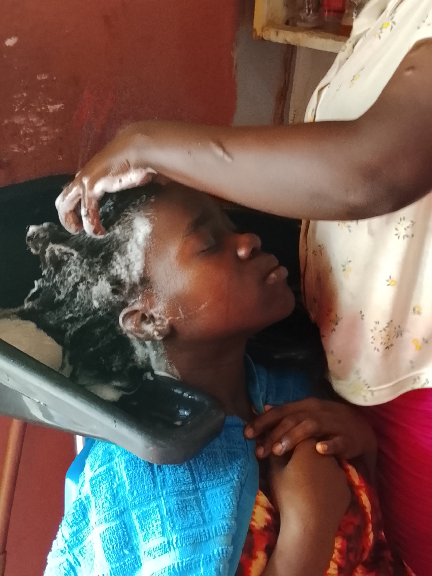 An indoor, eye-level shot captures a hair washing scene in Akwanga, Nigeria. The primary subject is a person, likely a young woman or girl, with dark skin, lying back in a black hair washing basin. Her eyes are closed, suggesting relaxation, while her dark, textured hair is thoroughly covered in white shampoo suds. A bright blue towel with a textured pattern is draped around her shoulders, and her hands are gently clasped over another multi-colored, patterned fabric (possibly another towel or part of her clothing) resting on her chest.
A second person, likely a stylist, is visible from the elbow down, with their dark-skinned hand actively massaging the shampoo into the first person's hair. This stylist wears a light-colored top with a subtle floral or leaf pattern and what appears to be a red skirt or trousers.
The background features a reddish-brown wall, giving the impression of an interior space, possibly a home or a local salon. In the upper right corner, a portion of a wooden shelf is visible, with faint outlines of objects, possibly bottles or containers. The overall atmosphere appears calm and focused on the hair care activity. No visible text is present in the image.