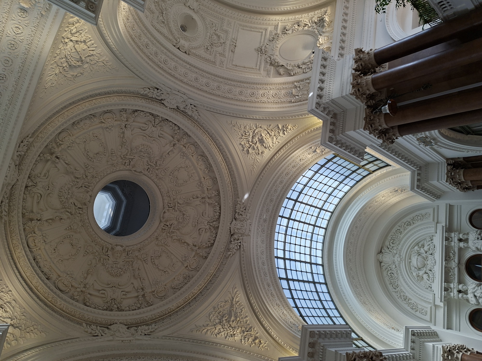The image shows an interior view of the Royal Galleries of Saint-Hubert in Brussels, Belgium. The scene is dominated by ornate architecture, featuring a high, domed ceiling with intricate plasterwork and decorative reliefs. A large, arched glass roof allows natural light to illuminate the space. In the foreground, massive Corinthian columns with reddish-brown shafts and ornate capitals support the upper structures. The overall impression is one of grandeur and classical design. There are no people visible in the image.