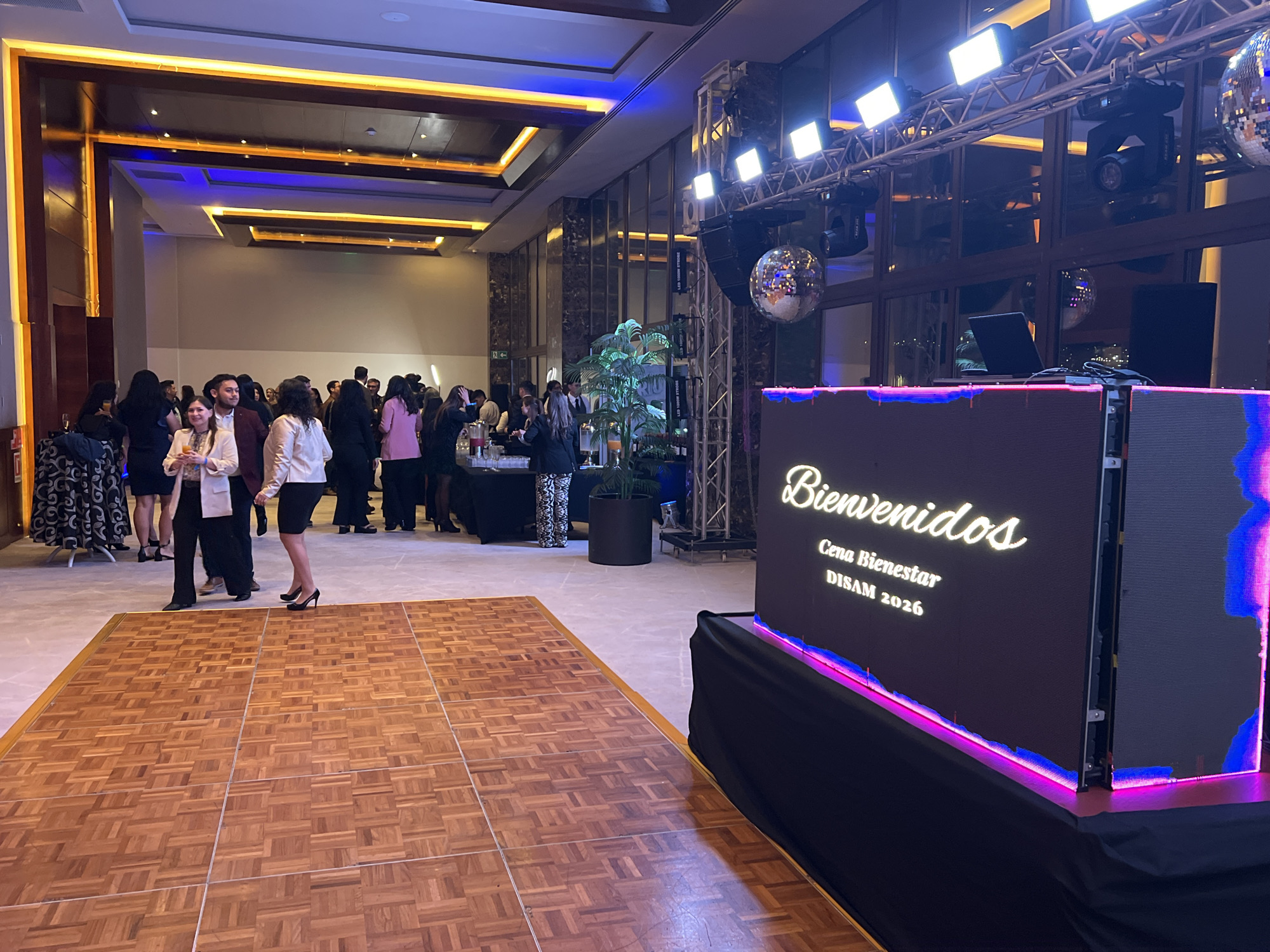 The image depicts an indoor event, possibly a formal gathering or party, set within a spacious hall. The environment features elegant interior design with prominent use of lighting, including illuminated ceiling accents in warm orange and cool blue tones. A dance floor, constructed from polished wooden tiles arranged in a parquet pattern, occupies the foreground. To the right, a large LED screen is set up, displaying "Bienvenidos" in a script font, followed by "Cena Bienestar DISAM 2026" in a plainer font. The screen is framed by vibrant pink and blue LED lights.
In the background, a crowd of formally dressed individuals, men and women, are mingling and standing around. Some appear to be holding drinks. There's a bar or buffet area visible on the left side of the image with bottles and serving containers. Further to the right, professional lighting equipment and a disco ball are suspended from a metal truss structure, suggesting a DJ or entertainment setup. The overall atmosphere suggests a celebratory or festive occasion. The image does not provide any specific information that would allow for identification of the location as Arica, Chile.