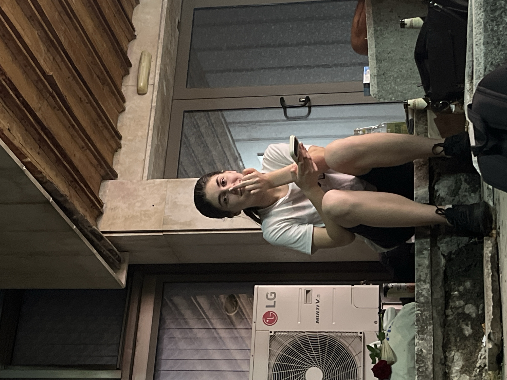 A young woman with dark hair pulled back in a ponytail is sitting on concrete steps outside a building. She is wearing a white t-shirt and dark shorts, with black sneakers on her feet. She is holding a smartphone in her left hand and has her right hand near her mouth. To her left is a white LG air conditioning unit. In front of the air conditioning unit, on the steps, is a red rose placed on a white plastic bag. Several beer bottles and a pack of cigarettes are visible on the steps behind her. The building has a wooden roof structure and large glass doors. The scene appears to be outdoors in an urban environment, possibly in the city of Vratsa, Bulgaria.