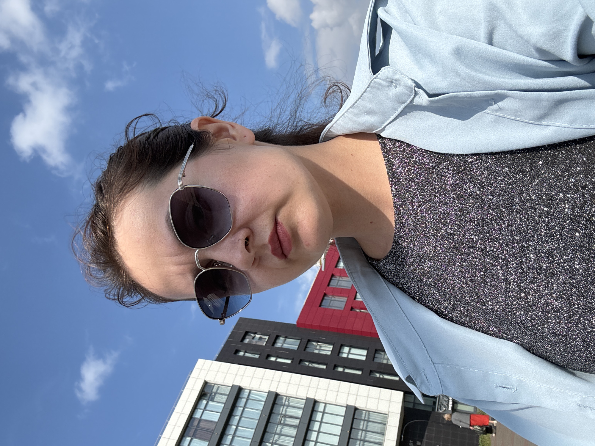 A woman wearing sunglasses and a light blue jacket over a sparkly dark top stands outdoors, looking at the camera. She has dark hair pulled back. Behind her are modern buildings with glass windows and a red accent wall, under a blue sky with some clouds. The scene appears to be in a city.