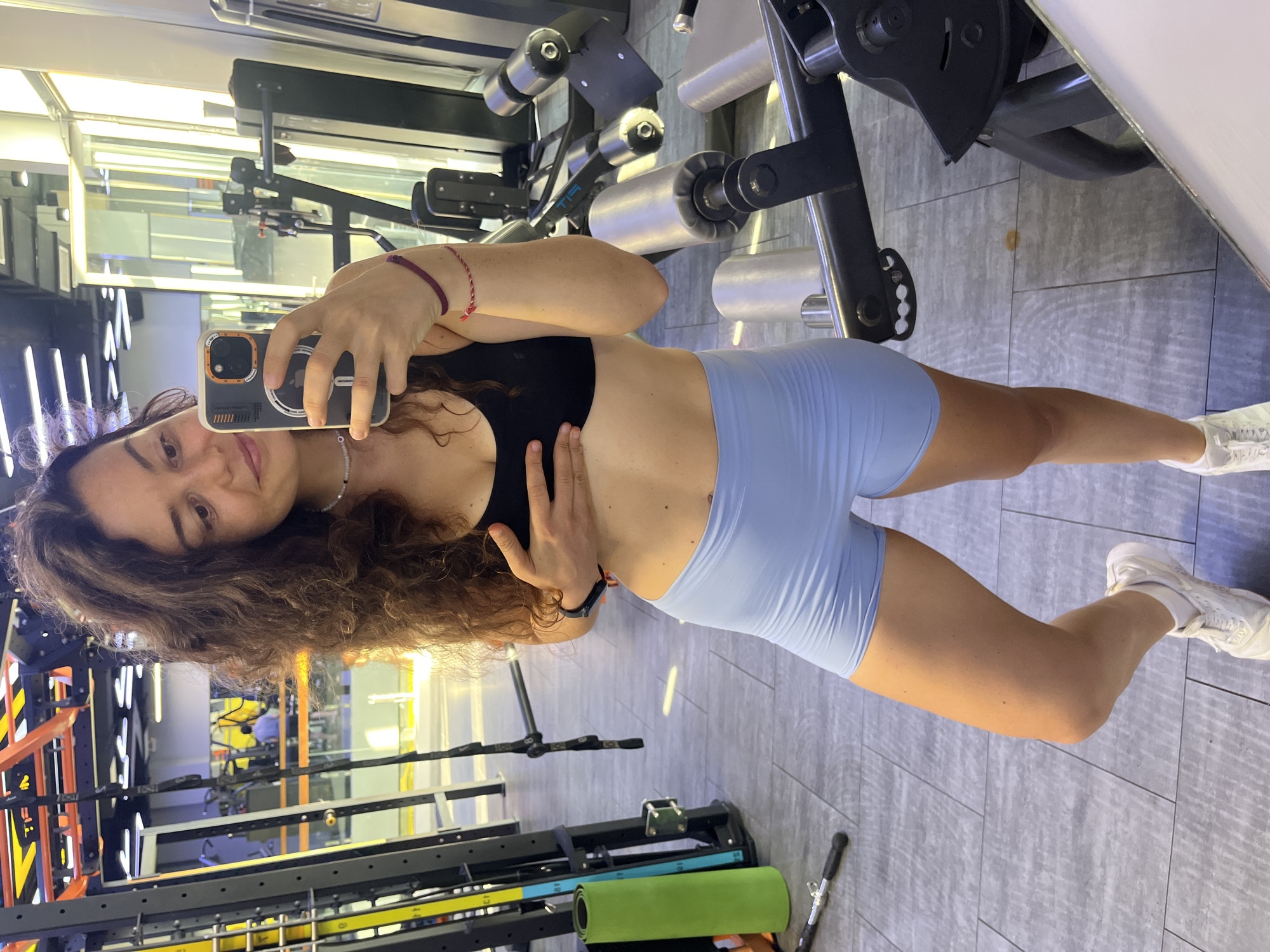 A young woman is taking a selfie in a gym. She is wearing a black sports bra and light blue athletic shorts. Her left hand is resting on her chest. She has curly brown hair and is wearing a necklace and two bracelets. In the background, there are gym machines and equipment. The flooring is tiled and the lighting suggests it is daytime. The image was taken in Phu Thuy Ward, Vietnam.