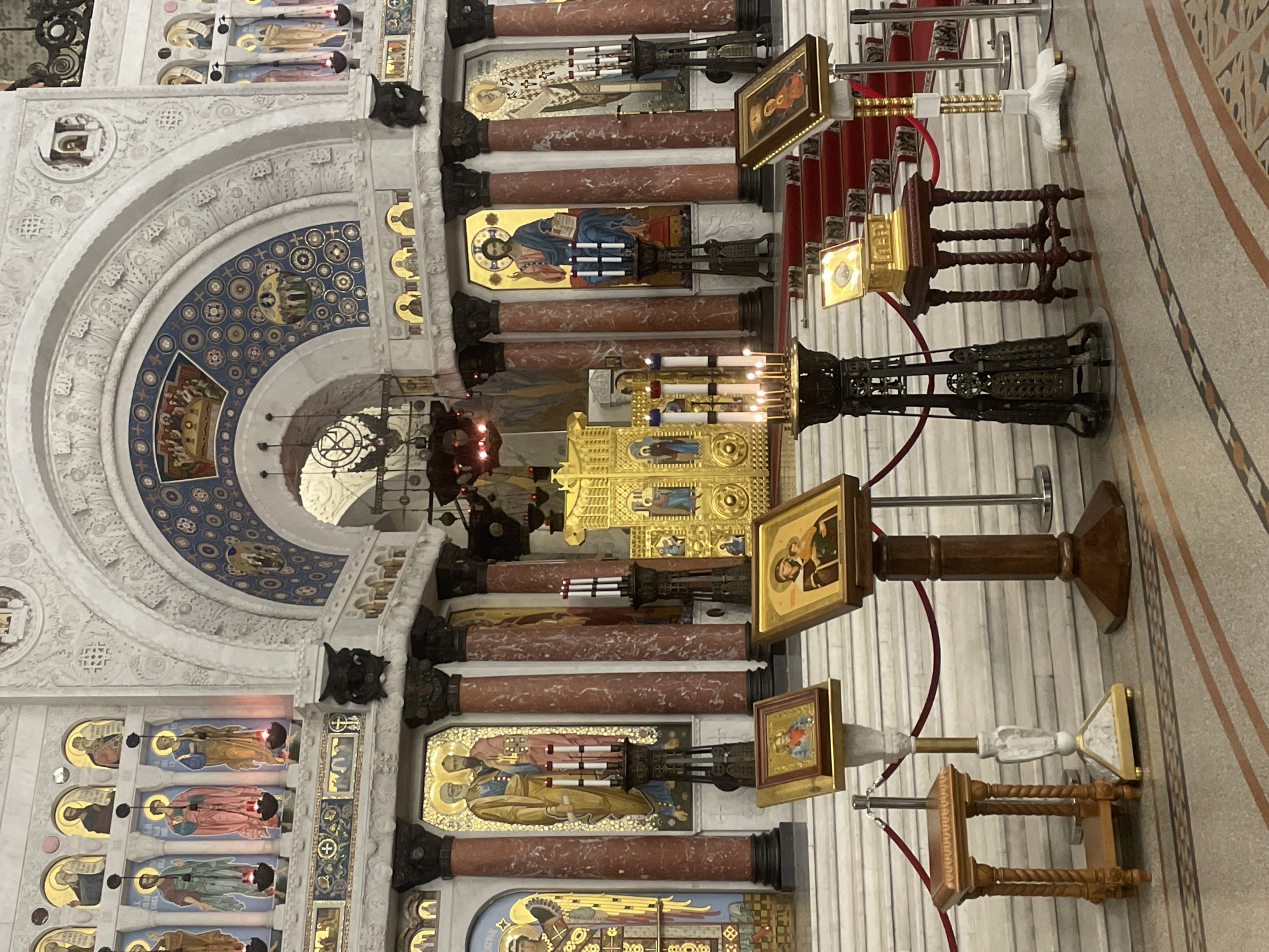 The image shows the interior of the Saint Nicholas Naval Cathedral in Kronstadt, Russia. The cathedral features ornate architectural details, including large marble columns, gilded iconostasis, and colorful murals depicting religious scenes. Several icons are displayed on stands in the foreground, with lit candles in front of them. The floor is a patterned mosaic. There are no people visible in the image.