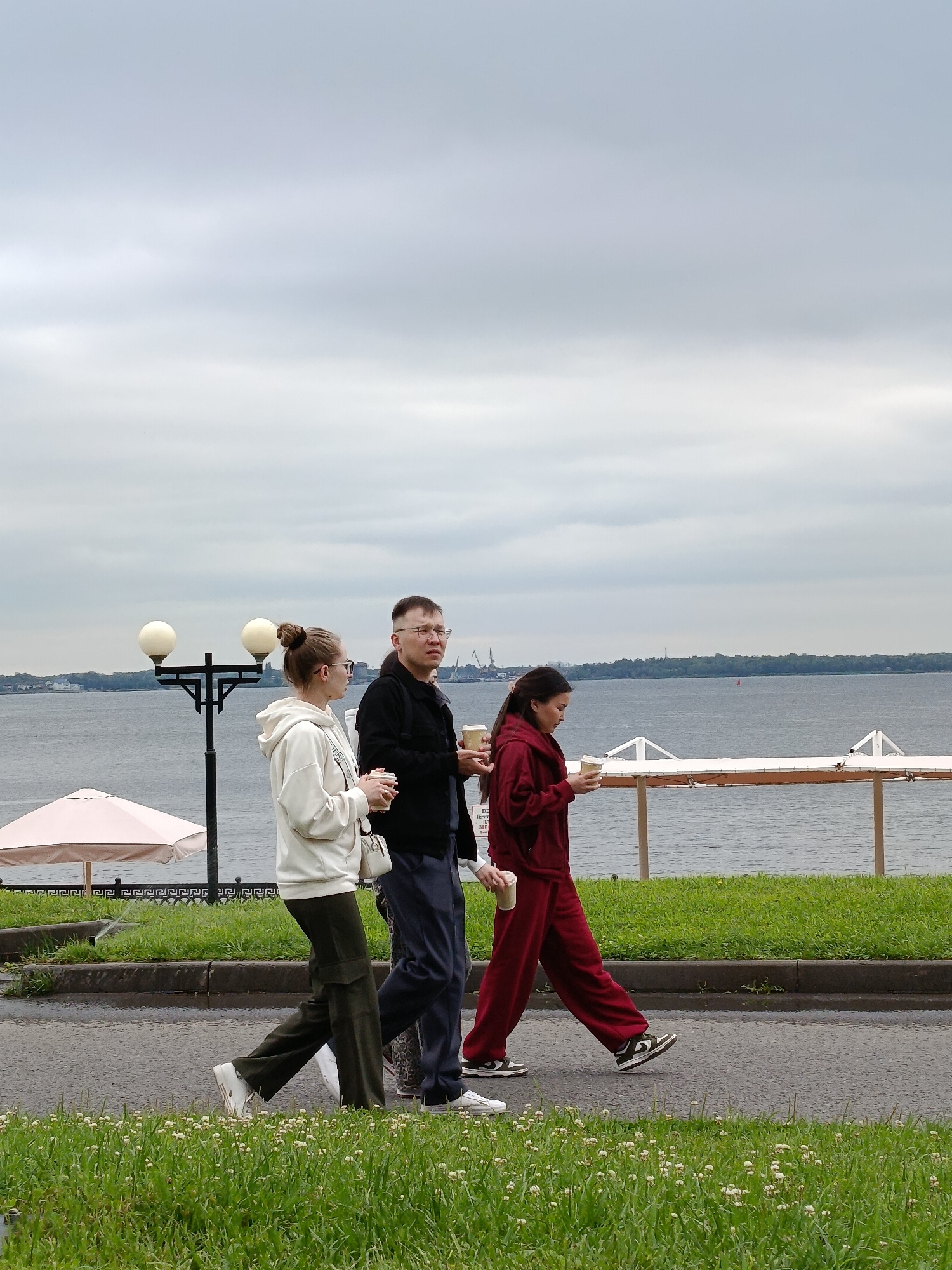 In Voskresensky District, Russia, three individuals are walking along a paved path by a large body of water under an overcast sky. On the left, a young woman with blonde hair tied in a bun, wearing glasses, a cream-colored hoodie, and dark green cargo pants, carries a paper cup. To her right, a man with glasses, wearing a black jacket and dark pants, also holds a paper cup. To the right of the man, a young woman with dark hair, wearing a maroon tracksuit, walks with a paper cup in her hand. In the background, a body of water stretches to the horizon, with trees lining the distant shore. A lamppost with two globes stands to the left of the group, and further left, a light pink umbrella is visible. A white fence with decorative elements runs along the edge of the water. The ground in the foreground is covered in green grass with small white flowers.