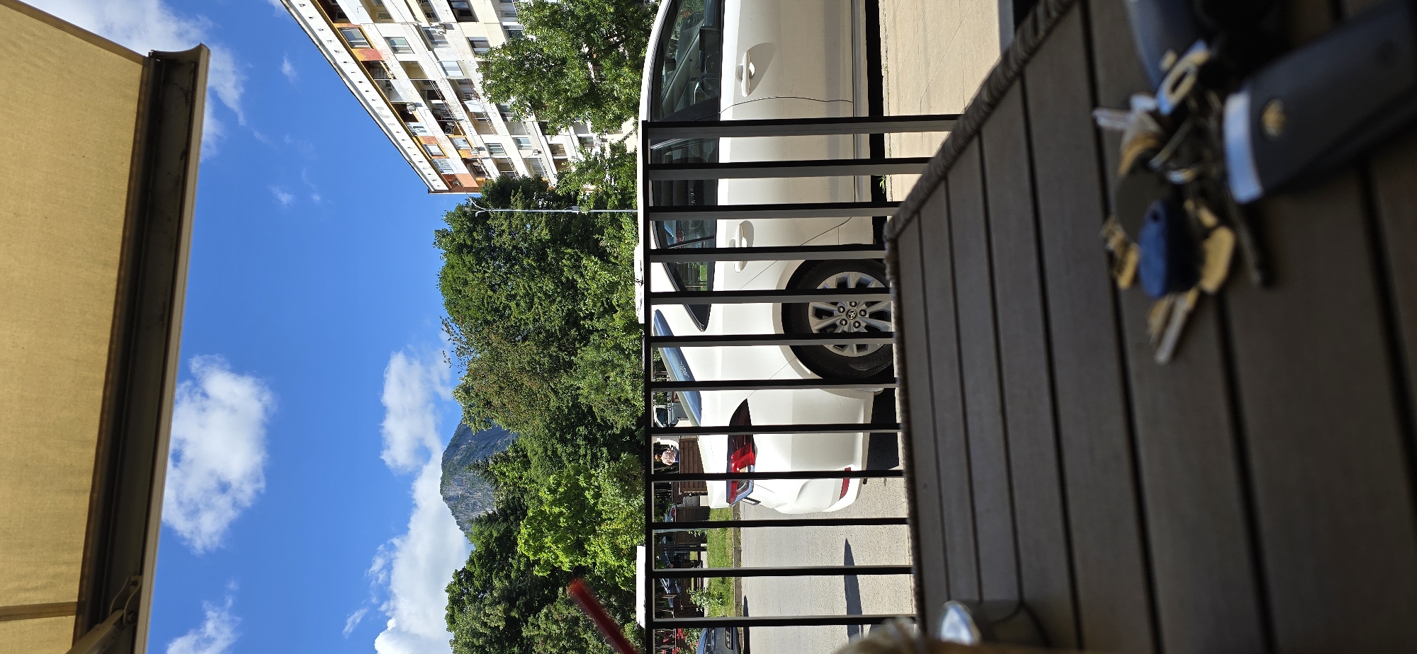 The image depicts an outdoor scene viewed from a seated perspective, with a wooden table in the foreground. On the table, a set of keys, including a car key fob with a blue tag, is scattered. Beyond the table, a black metal railing separates the viewer from a street scene. A white sedan is parked on the street, its rear section and side visible. The street is lined with lush green trees, and in the background, a mountain peak rises against a bright blue sky with scattered white clouds. To the right of the trees and mountain, a multi-story apartment building with white and orange accents is visible. The overall environment suggests a sunny day in a city with natural elements.