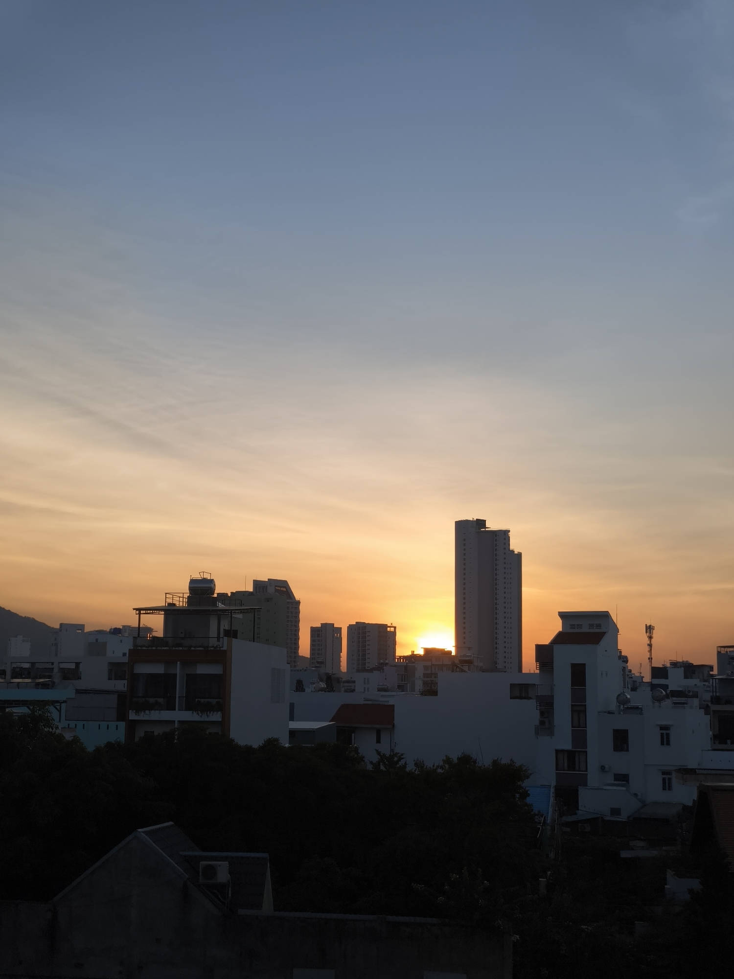 The image depicts a city skyline at sunset. The sky transitions from a pale blue at the top to a warm orange hue near the horizon, with wispy clouds adding texture. Several buildings of varying heights are visible, some appearing as silhouettes against the vibrant sky. In the foreground, darker shapes suggest trees and lower structures, possibly houses or smaller buildings. The overall scene captures a moment of natural beauty in an urban setting. The location is identified as Unknown, Unknown.