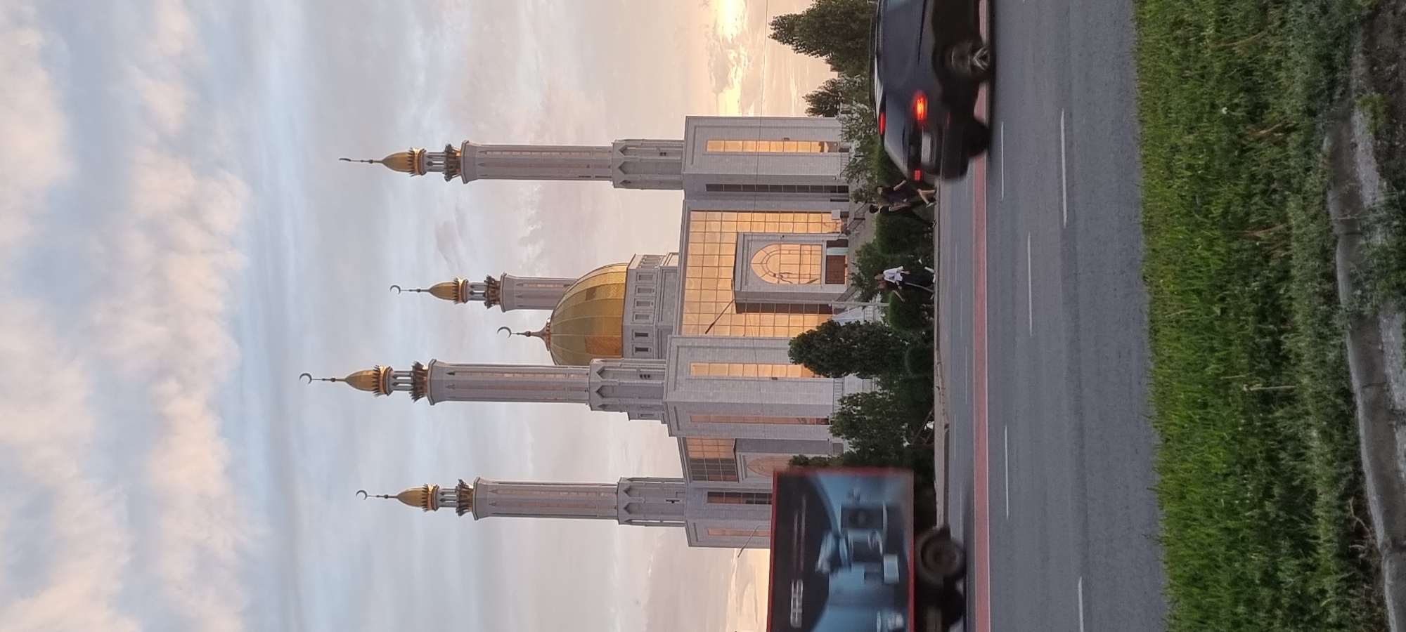 The image shows the Nur-Gasyr mosque in Aktobe, Kazakhstan, on a cloudy day. The mosque features a large golden dome and four tall minarets, each topped with a crescent moon. The building's facade is made of light-colored stone and large glass windows that reflect the sky. In the foreground, a road with a red lane marker runs horizontally across the frame. A blurred truck with an advertisement on its side is moving from left to right. Further down the road, a black SUV is also in motion, its taillights illuminated. Several people are walking on the sidewalk in front of the mosque, amidst green bushes and trees. The sky is filled with soft, diffused clouds, indicating either early morning or late afternoon light.