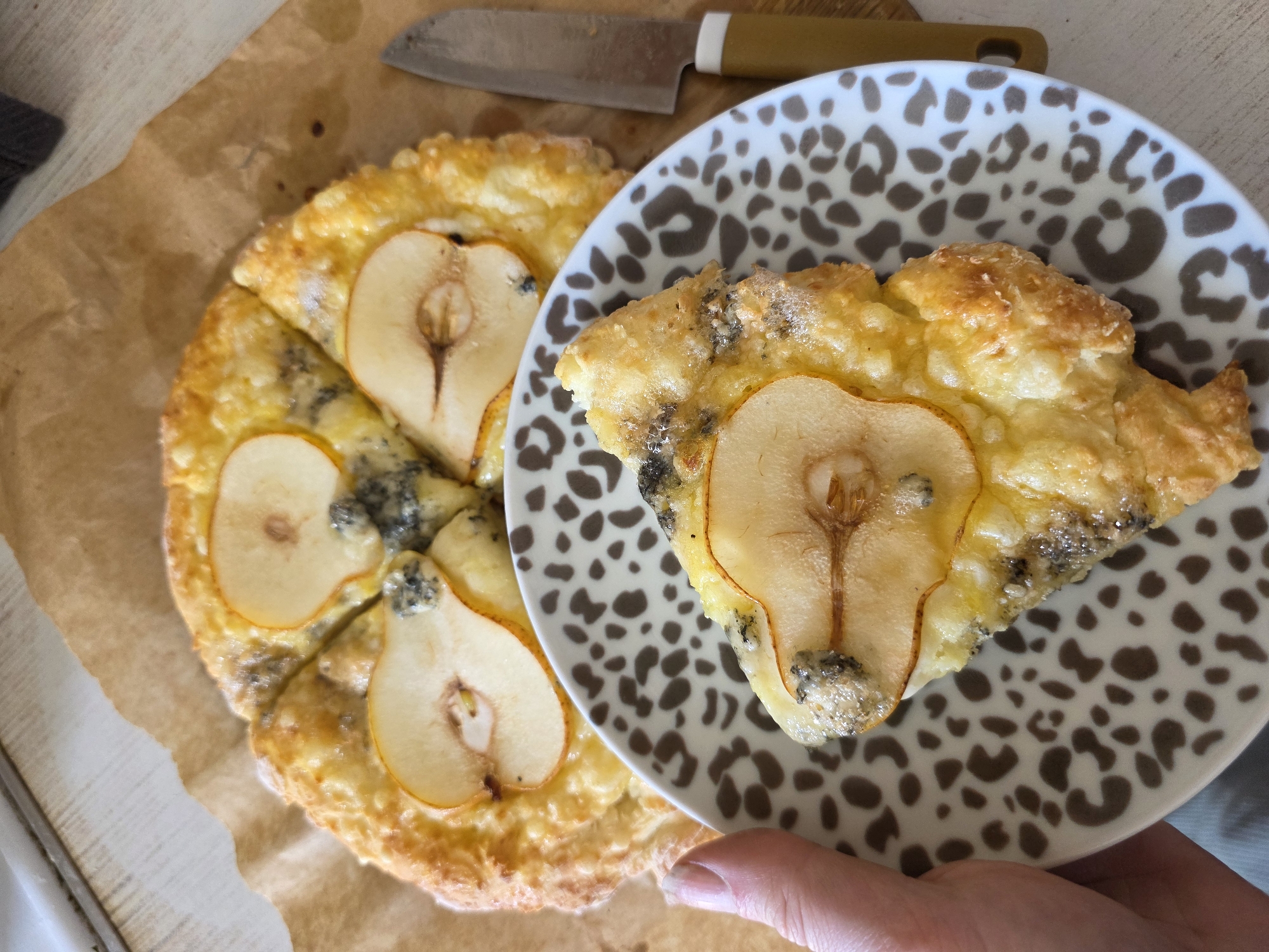 The image depicts a pear and cheese tart, with a slice on a small plate with a leopard print design. The tart is golden brown and the pears are pale with dark brown seeds. The cheese has a blue-grey hue. The plate is being held by a person. A knife with a beige handle is placed near the tart on a piece of brown parchment paper. The scene is indoors, likely on a kitchen counter or table. There is no evidence to suggest the location is Saint Petersburg, Russia.
