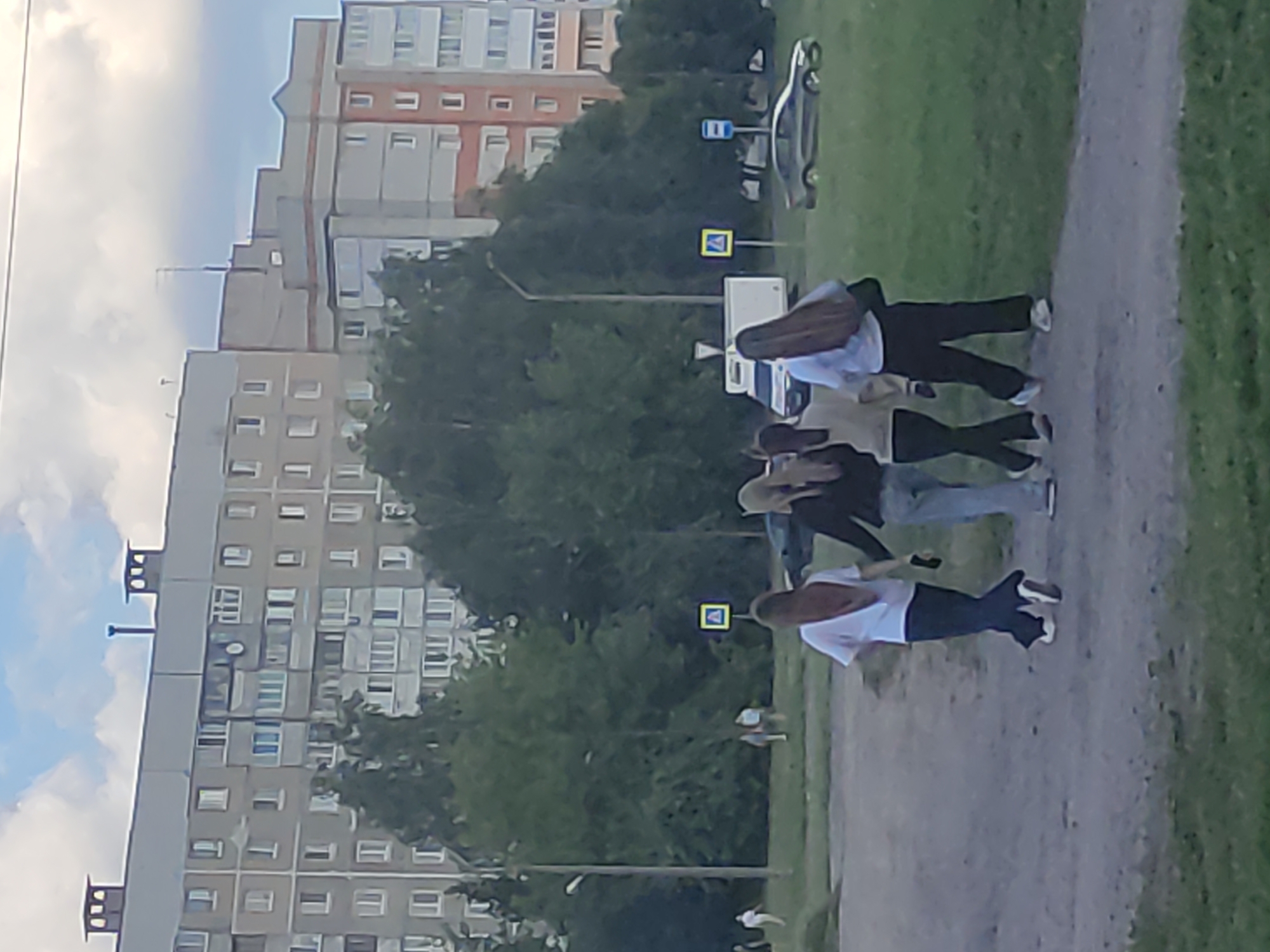 Four young women walk away from the camera on a dirt path, with grassy areas on either side. The woman on the far left wears a white t-shirt and dark pants. Next to her, a woman in a black jacket and light-colored pants walks with her. The third woman from the left is in a light-colored jacket and dark pants. The woman on the far right wears a dark jacket and dark pants. Behind them, a dark-colored sedan is parked on the grass. Across the path, a yellow pedestrian crossing sign is visible. In the background, several multi-story apartment buildings, typical of Soviet-era architecture, dominate the skyline, along with numerous trees. A street light is visible on the left side of the image. The sky is partly cloudy. The scene appears to be in an urban residential area in Mahilyow, Belarus.