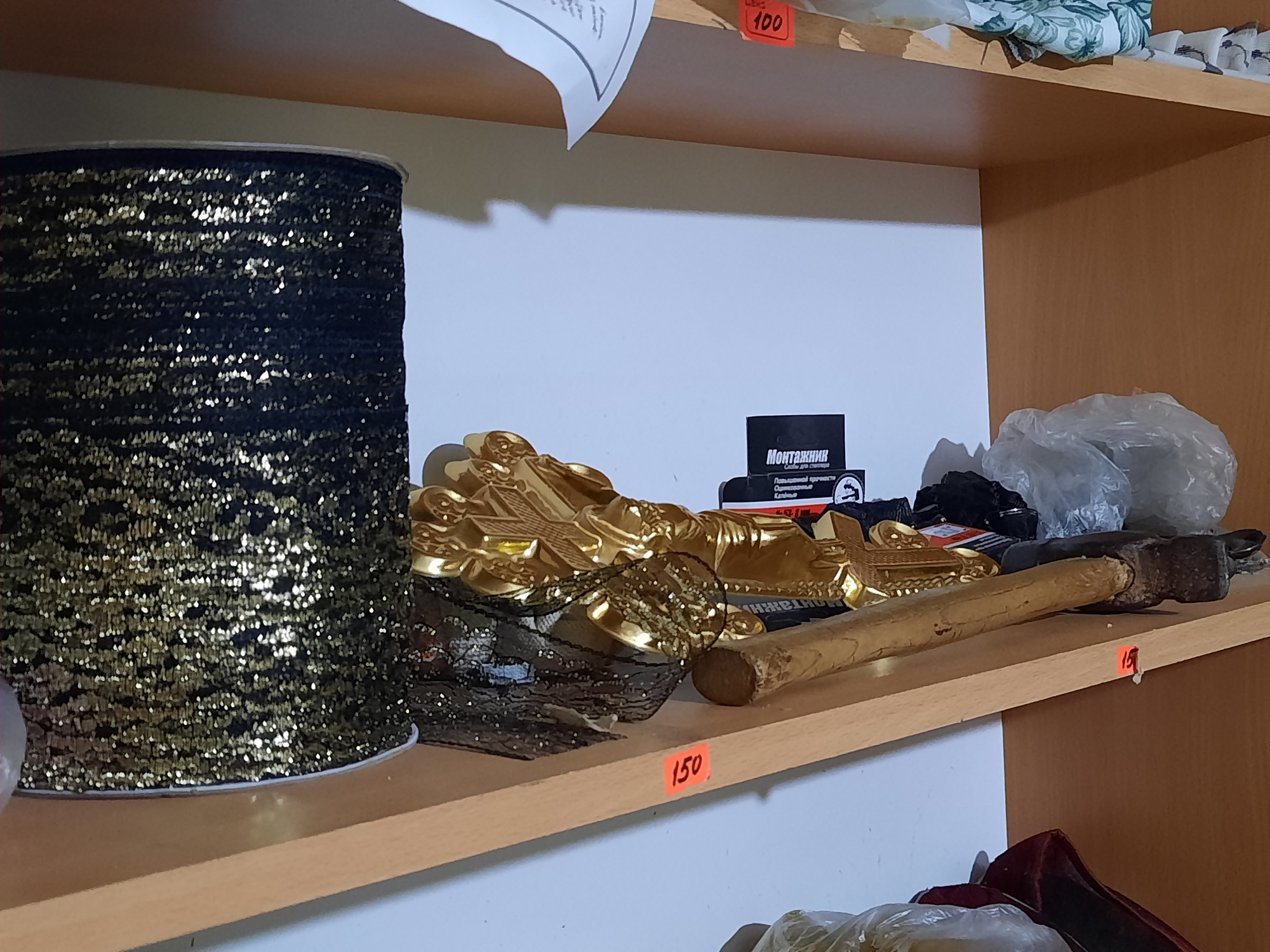 The image shows a cluttered shelf. On the shelf are rolls of black and gold ribbon, a decorative golden ornament, a black box with white lettering, a hammer, and plastic bags containing unseen contents. Price tags labeled "150" and "19" are visible on the shelf's edge. The location is likely a storage or shop area in Krasnogorskoye, Russia, based on the Cyrillic writing on the black box.