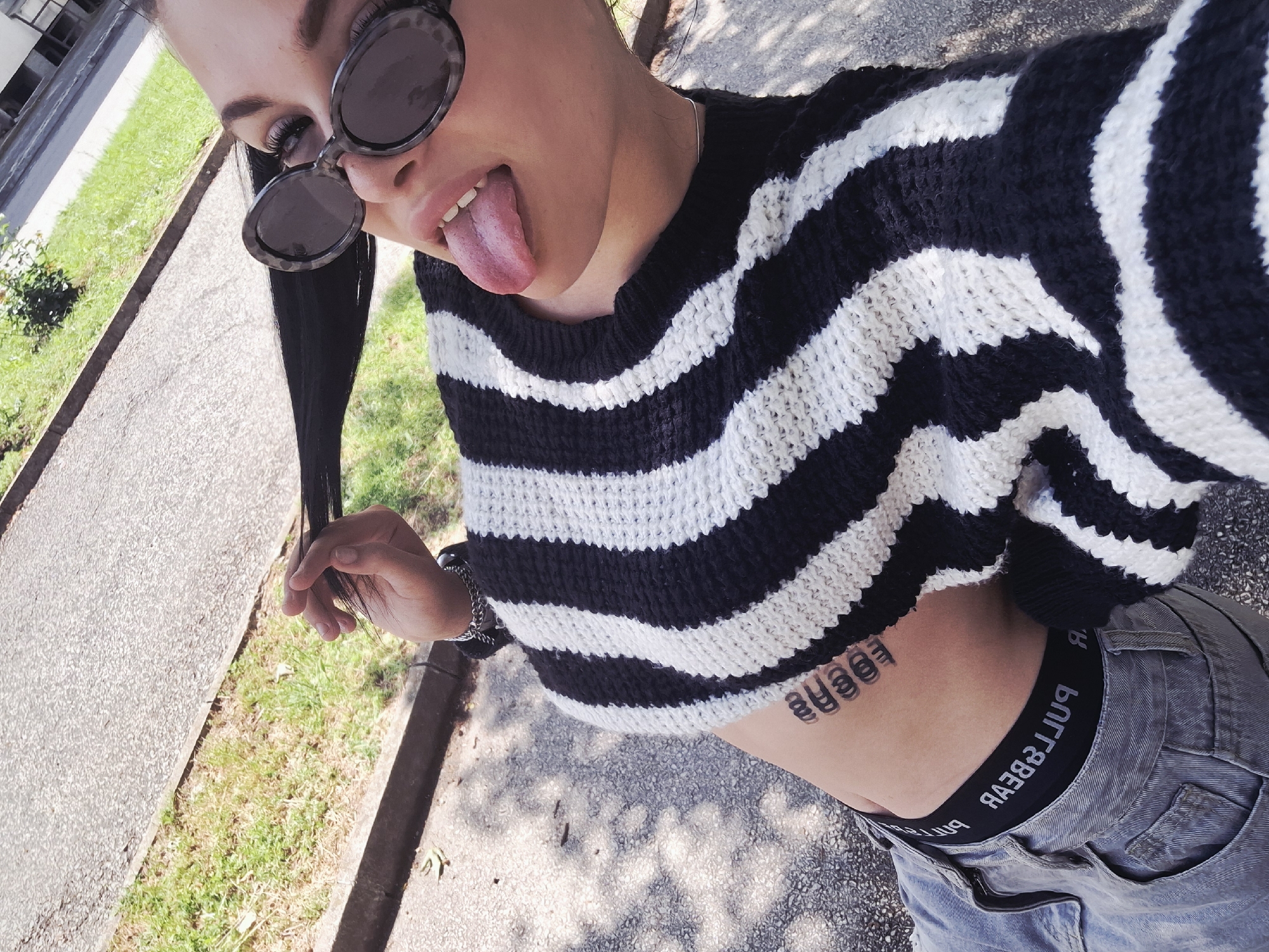 A young woman is captured in a playful outdoor selfie, looking down at the camera with her tongue sticking out. She has dark hair, a section of which she is holding with her left hand. Round, tortoiseshell-patterned sunglasses are perched on her head. She is wearing a black and white striped cropped knit sweater, exposing her midriff, and light gray jeans. A black underwear band is visible above her jeans, displaying the mirrored text "RA3B&LLUЯ," which reads "PULL&BEAR." A stylized tattoo of repeating circular or oval shapes is visible on her right ribcage. A silver chain bracelet adorns her left wrist. The setting appears to be outdoors in Mezdra, Bulgaria, under bright sunlight, indicated by visible shadows on the ground. The background features a paved path or sidewalk on the left and a grassy area with some green foliage on the right.