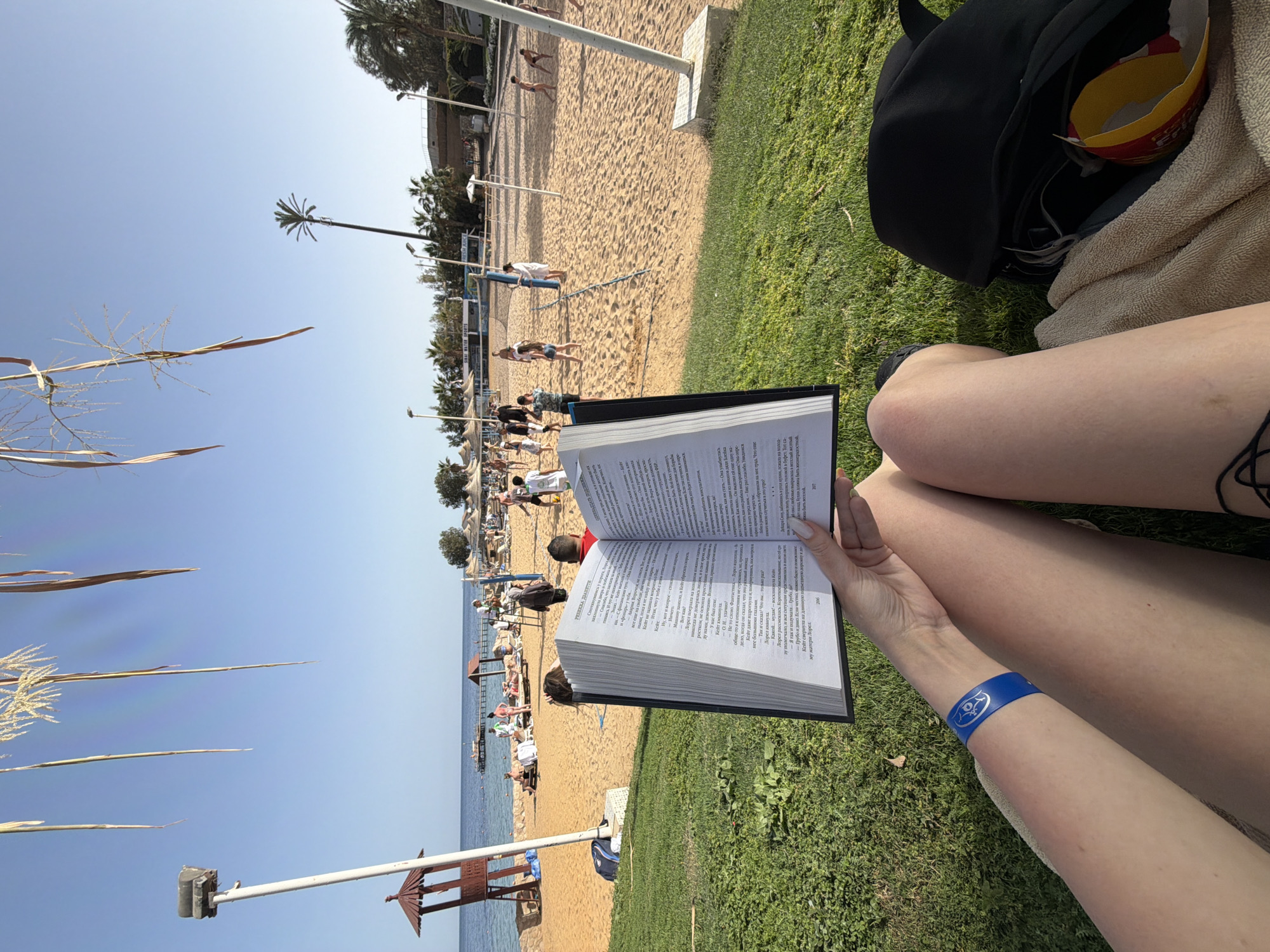 The image depicts a first-person perspective of someone sitting on grass near a sandy beach. The beach is populated with people and beach umbrellas, leading to the ocean. A person is holding an open book in their hands, with the pages displaying text in Russian. The person has a blue wristband on their left arm. In the background, there is a black backpack and a cardboard container for food. The location is likely Sharm El Sheikh, Egypt, based on the beach landscape.