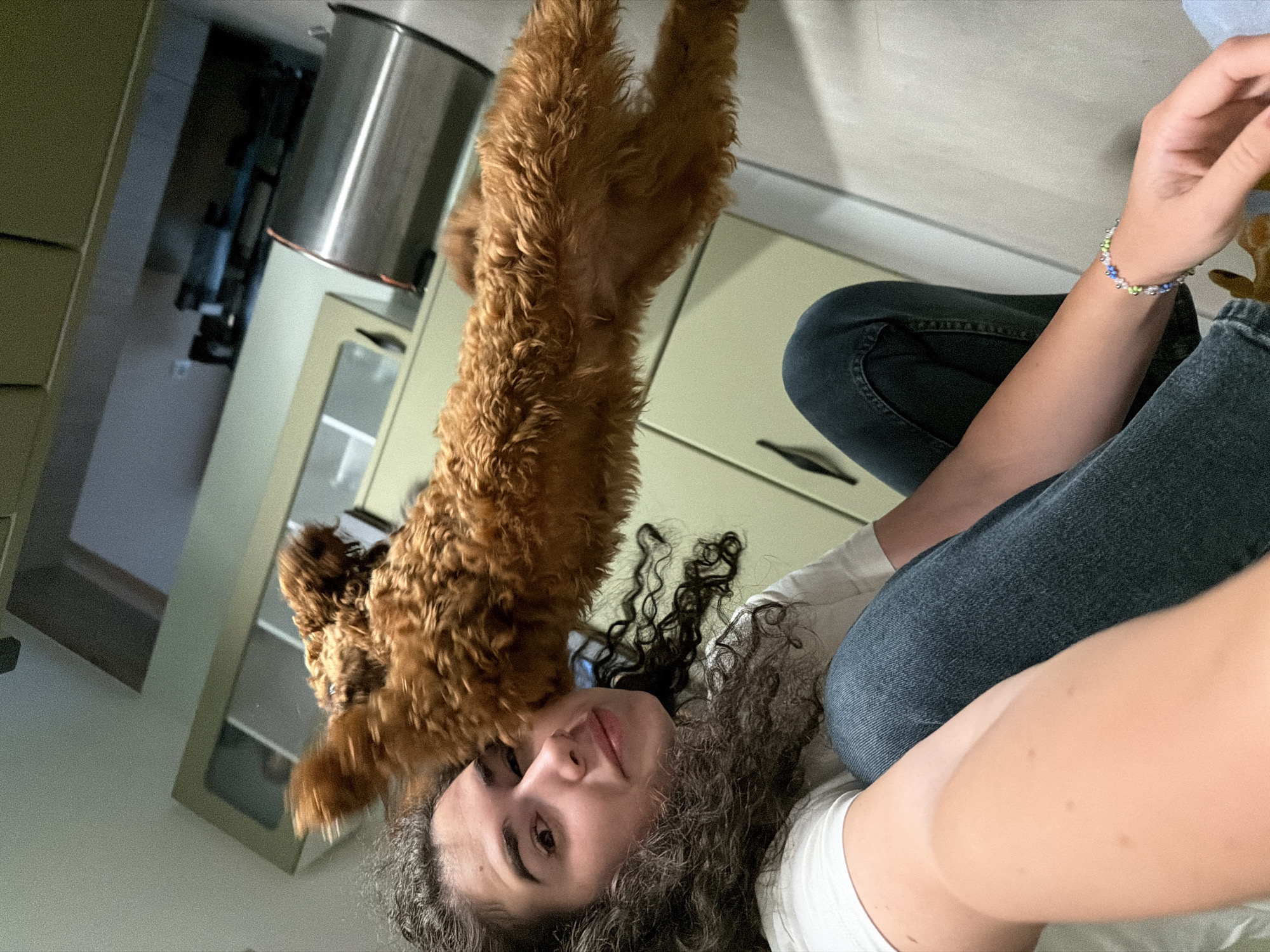 An overhead shot captures a young woman with curly dark hair, wearing a white t-shirt and denim jeans, crouched down. She is looking at a small, fluffy brown dog, a poodle mix, which is standing on its hind legs and reaching towards her. The dog's paws are blurred, indicating movement. The woman's left arm is bent, and her hand, adorned with a colorful beaded bracelet, is extended towards the dog. The background features kitchen cabinets, a silver trash can, and part of a doorway. The flooring appears to be linoleum or tile. The lighting is dim, suggesting an indoor setting. The scene is devoid of any identifiable landmarks or contextual cues that would place it within Sofia, Bulgaria.